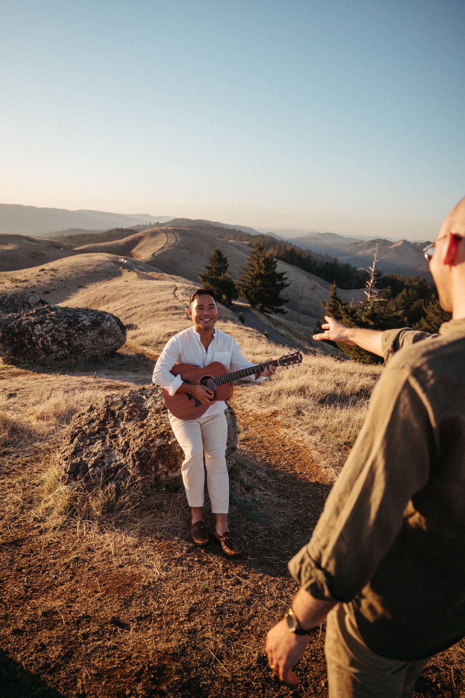 A smiling musician plays acoustic guitar on a sunny mountaintop while a companion gestures toward them.