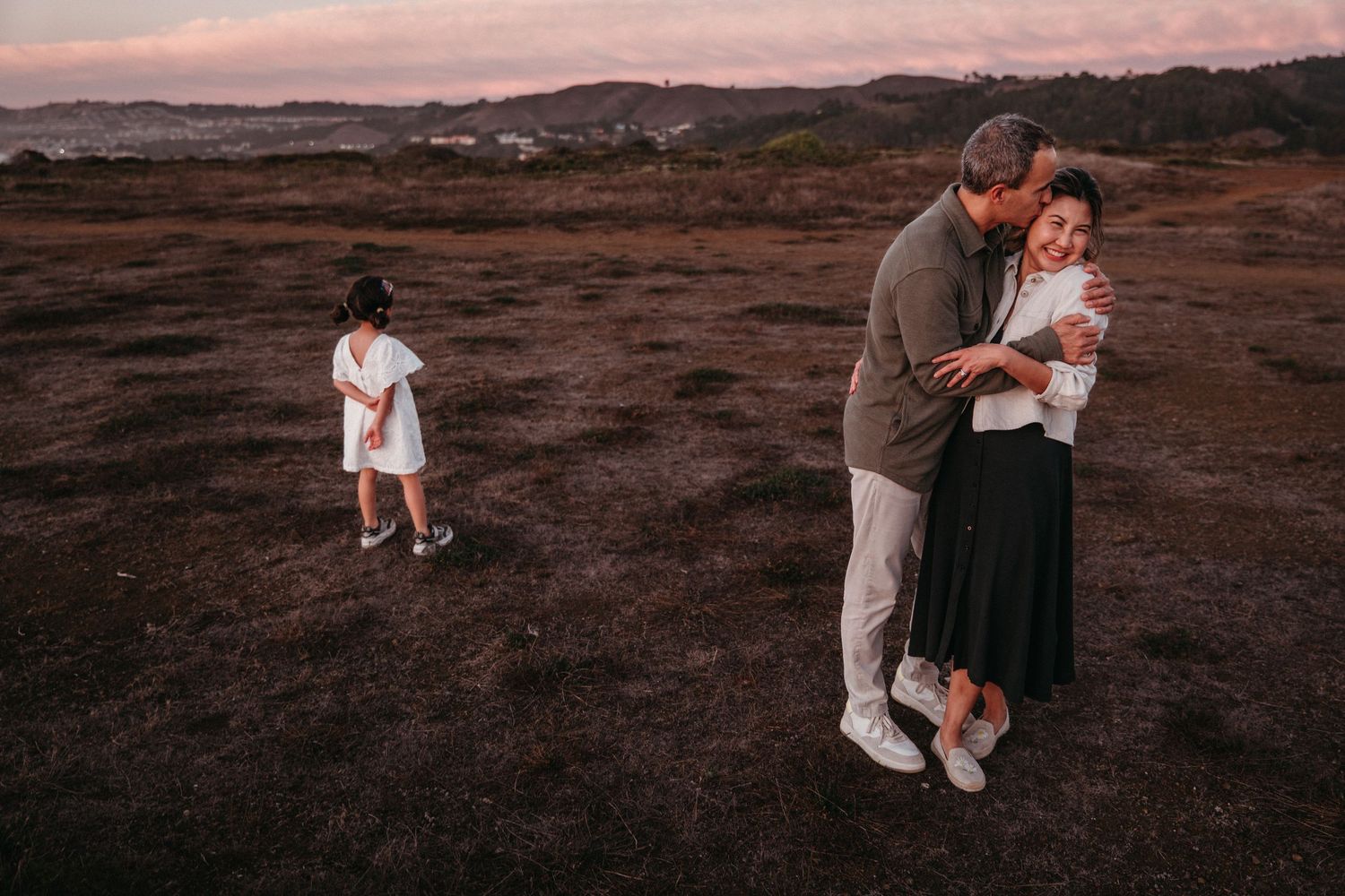 A couple embraces at dusk in an open field while a young girl stands nearby, with rolling hills in the background.