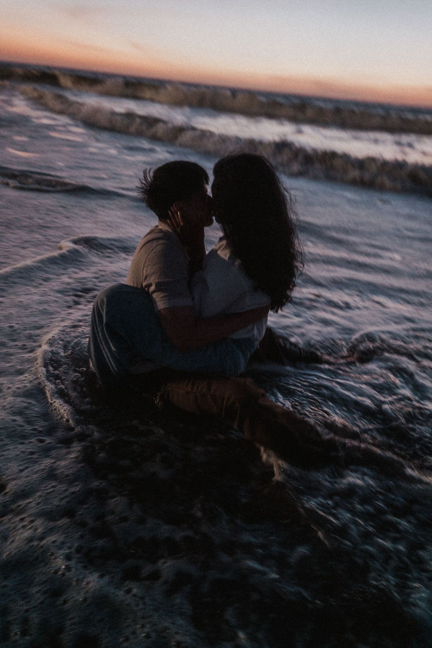 Romantic couple embracing and kissing while sitting in ocean waves on a beach at sunset.