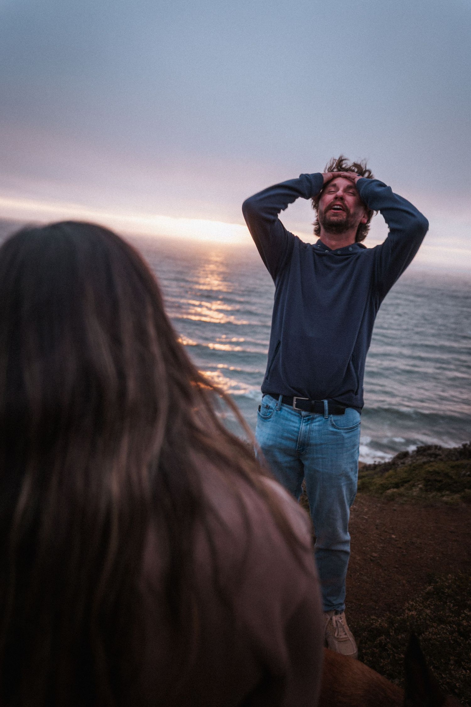 Man with hands on head stands at coastal cliff edge during sunset with ocean waves behind him.
