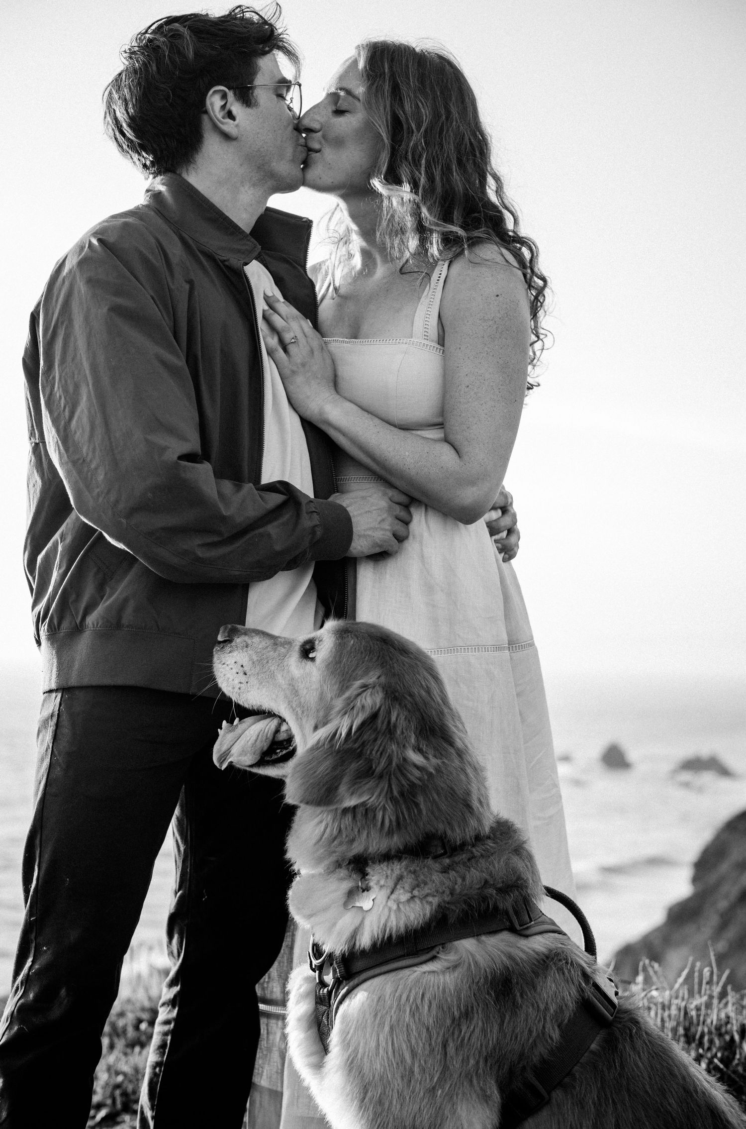 Black and white photo of a couple kissing outdoors by the ocean with their golden retriever dog sitting in front of them.