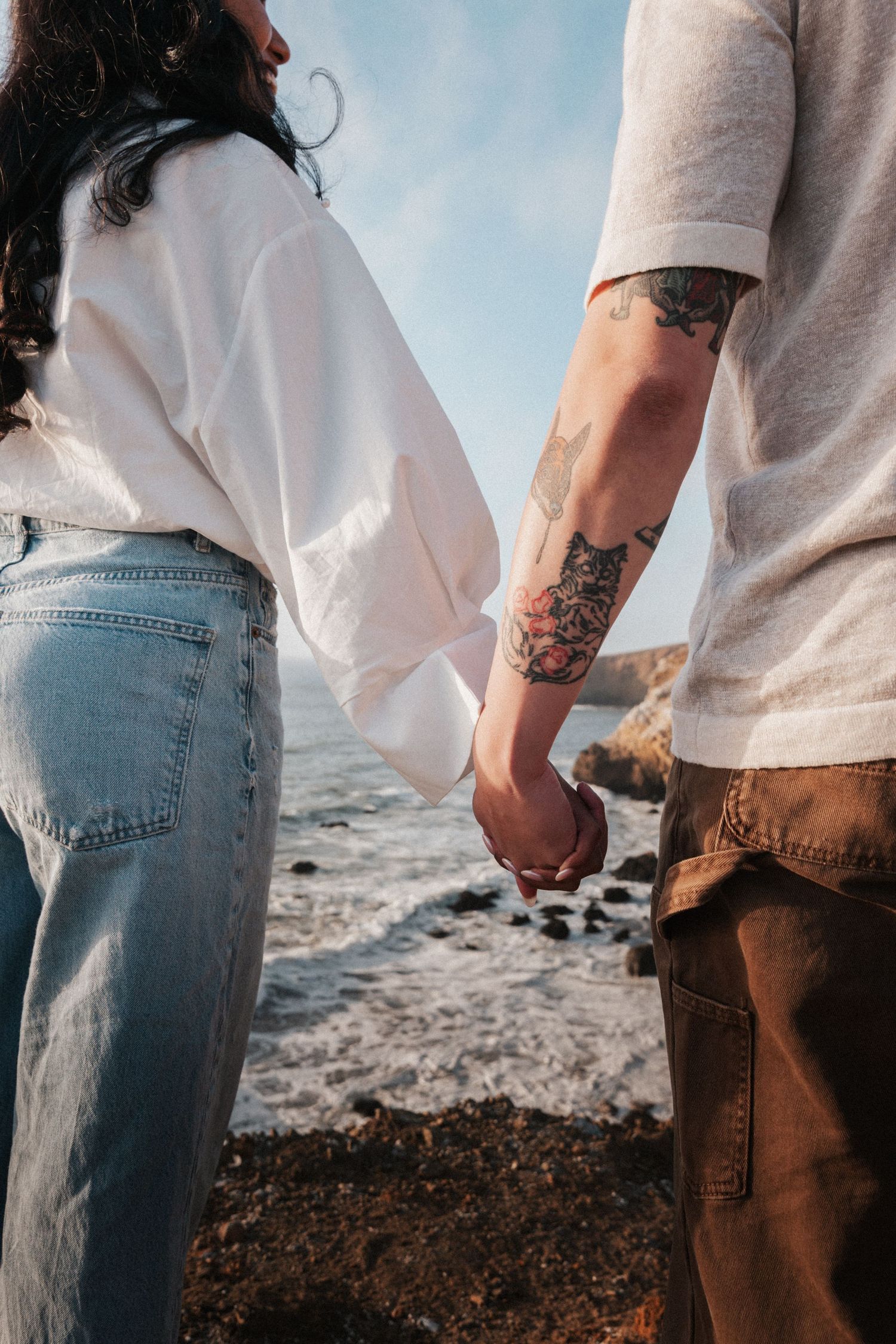 Close-up of a couple holding hands by the ocean, man with floral tattoo on forearm wearing white shirt.