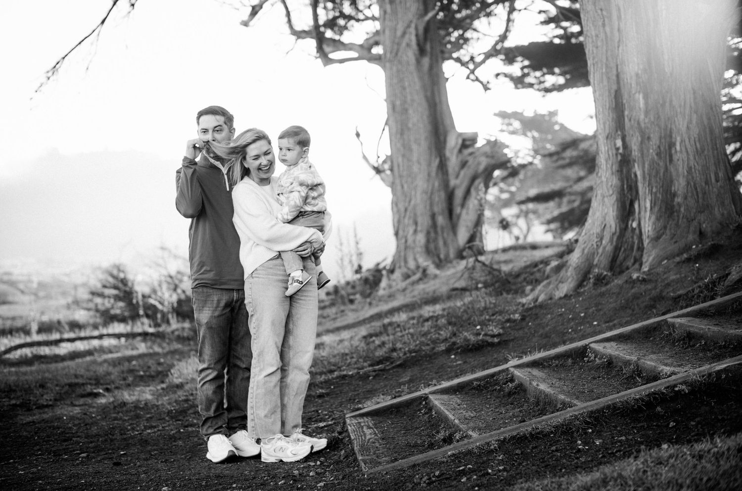 Black and white family photo of couple holding toddler outdoors near large trees in a park setting.