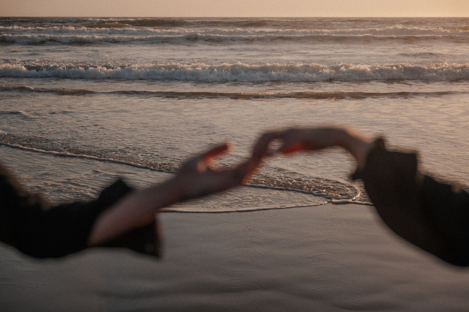 Two hands forming a heart shape together on a serene beach at sunset with gentle ocean waves in the background.