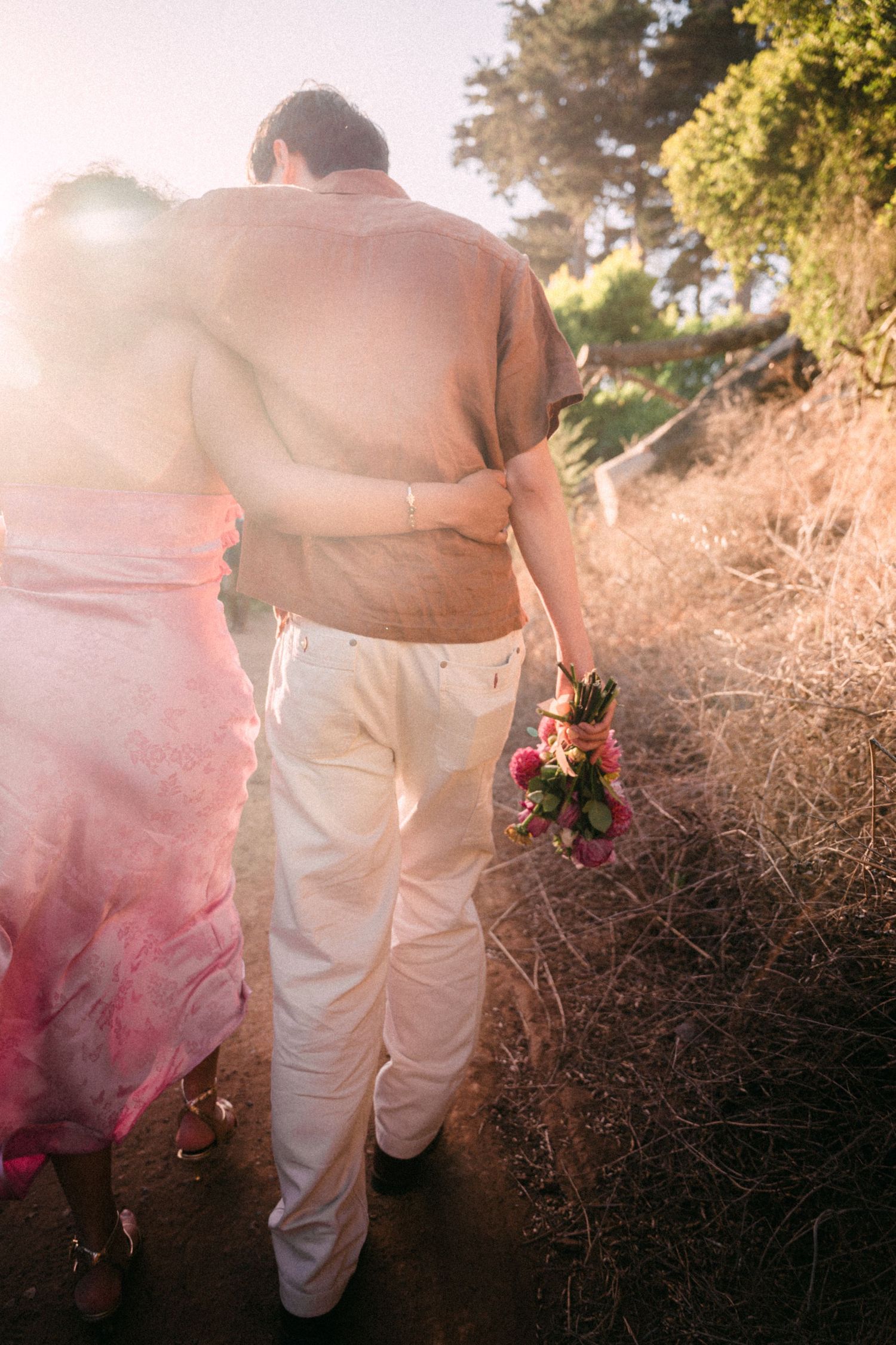 Couple walking on a sunlit path, man in white pants holding pink flowers with arm around woman in pink dress.