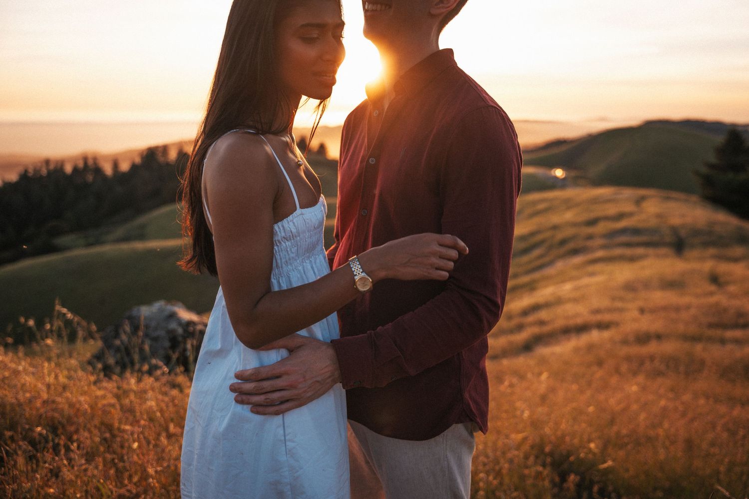 Couple embracing at golden sunset on a green hillside, woman in white dress, man in burgundy shirt.Photography by The Y.K. Studio.