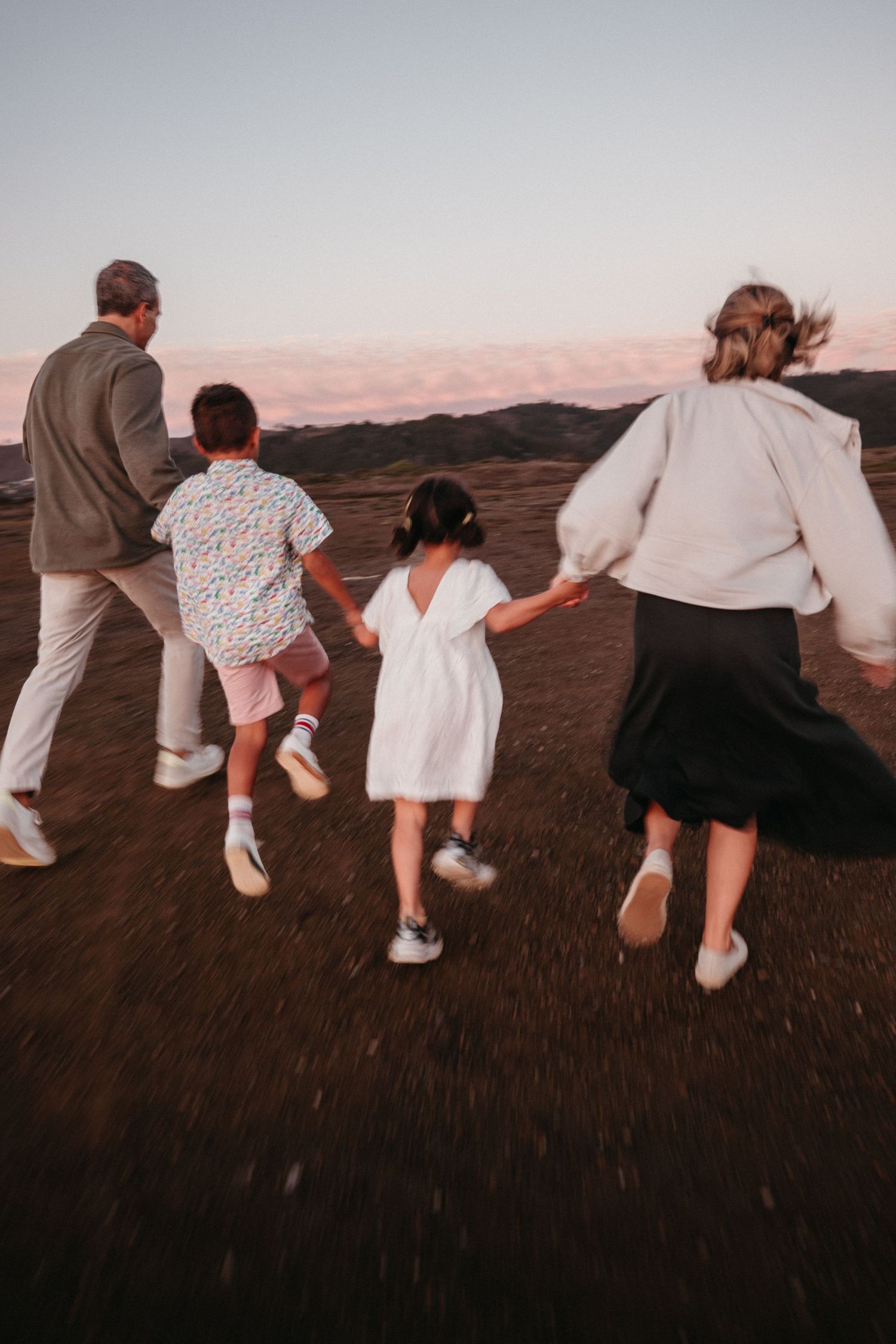 Family of four running hand-in-hand across a hilltop at sunset with mountains in the background.