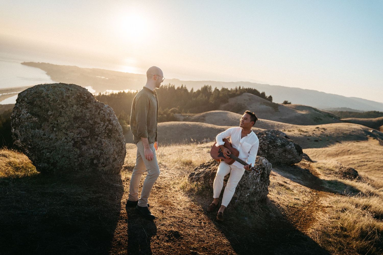Couple enjoying a scenic hilltop sunset view, one sitting on a rock, with a coastal landscape in the background.