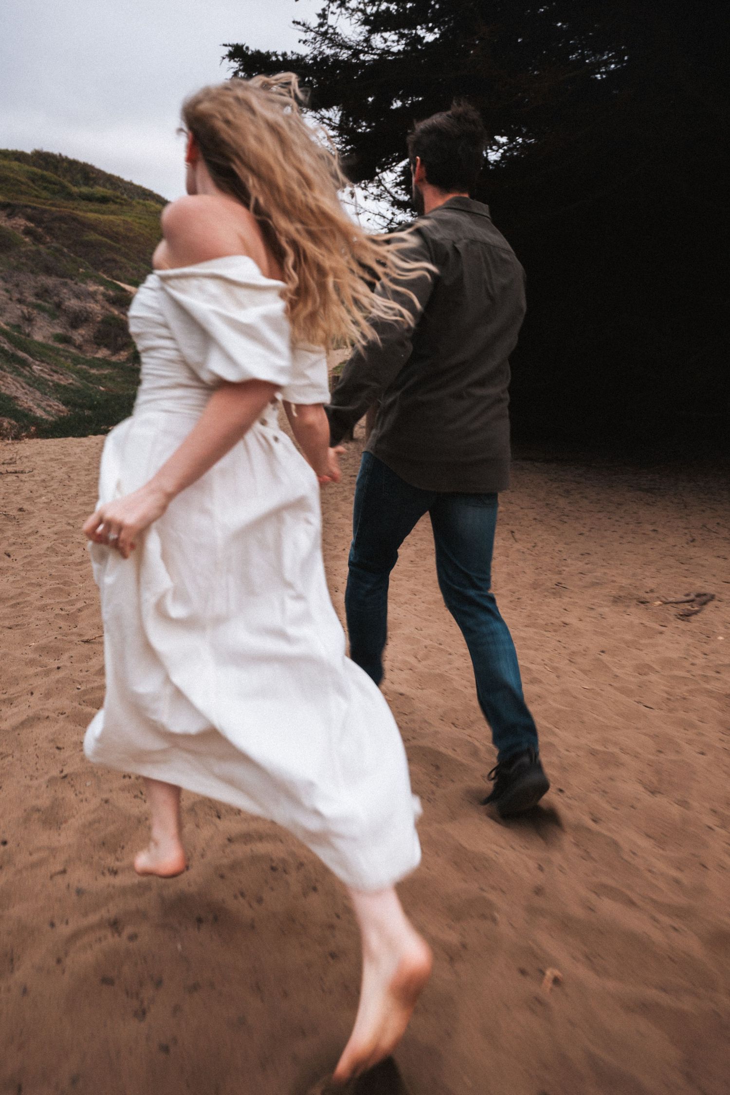 A couple holds hands running barefoot on a sandy beach, the woman in a white dress with wind-blown hair.