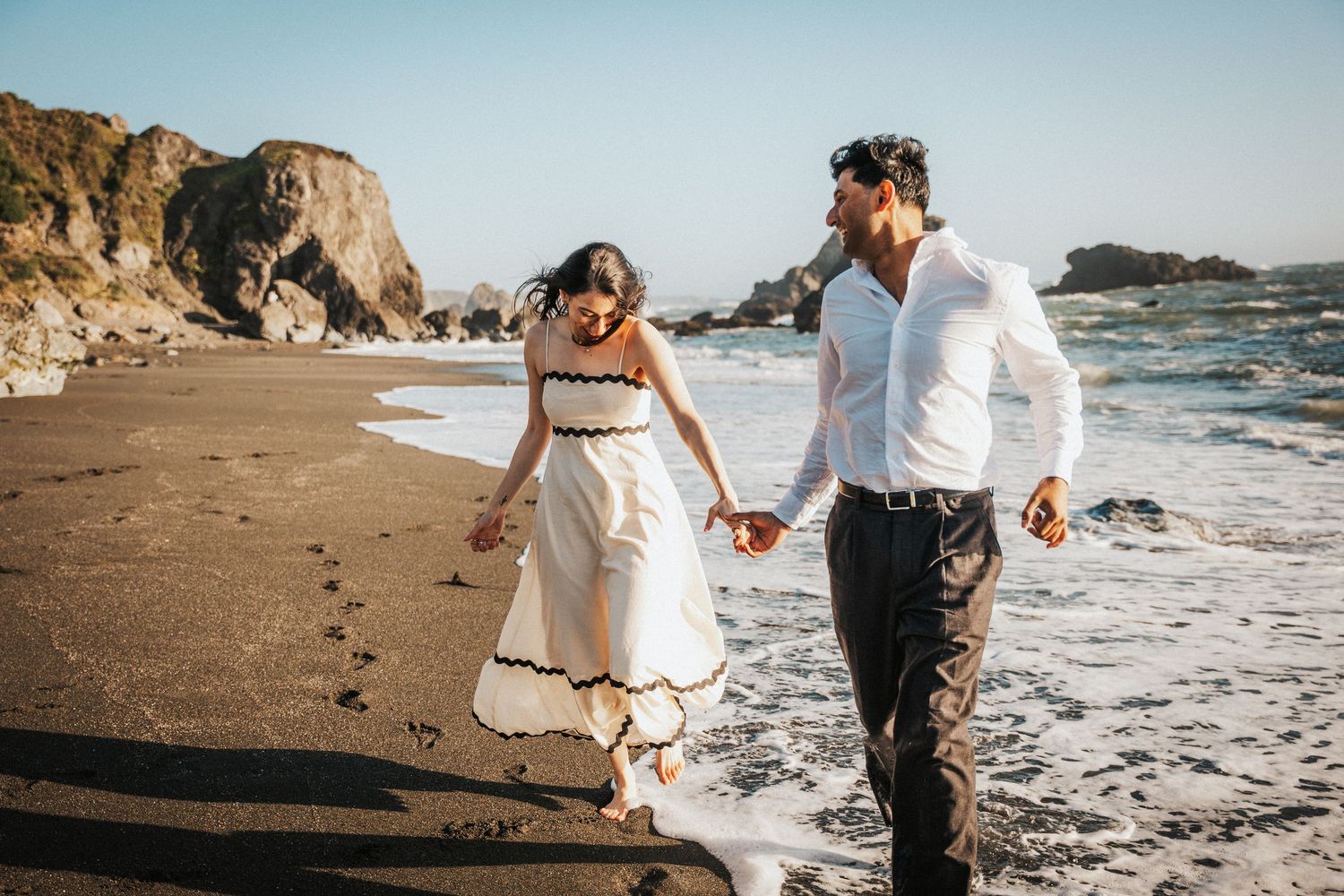 Couple holding hands walking along a black sand beach with ocean waves at golden hour.