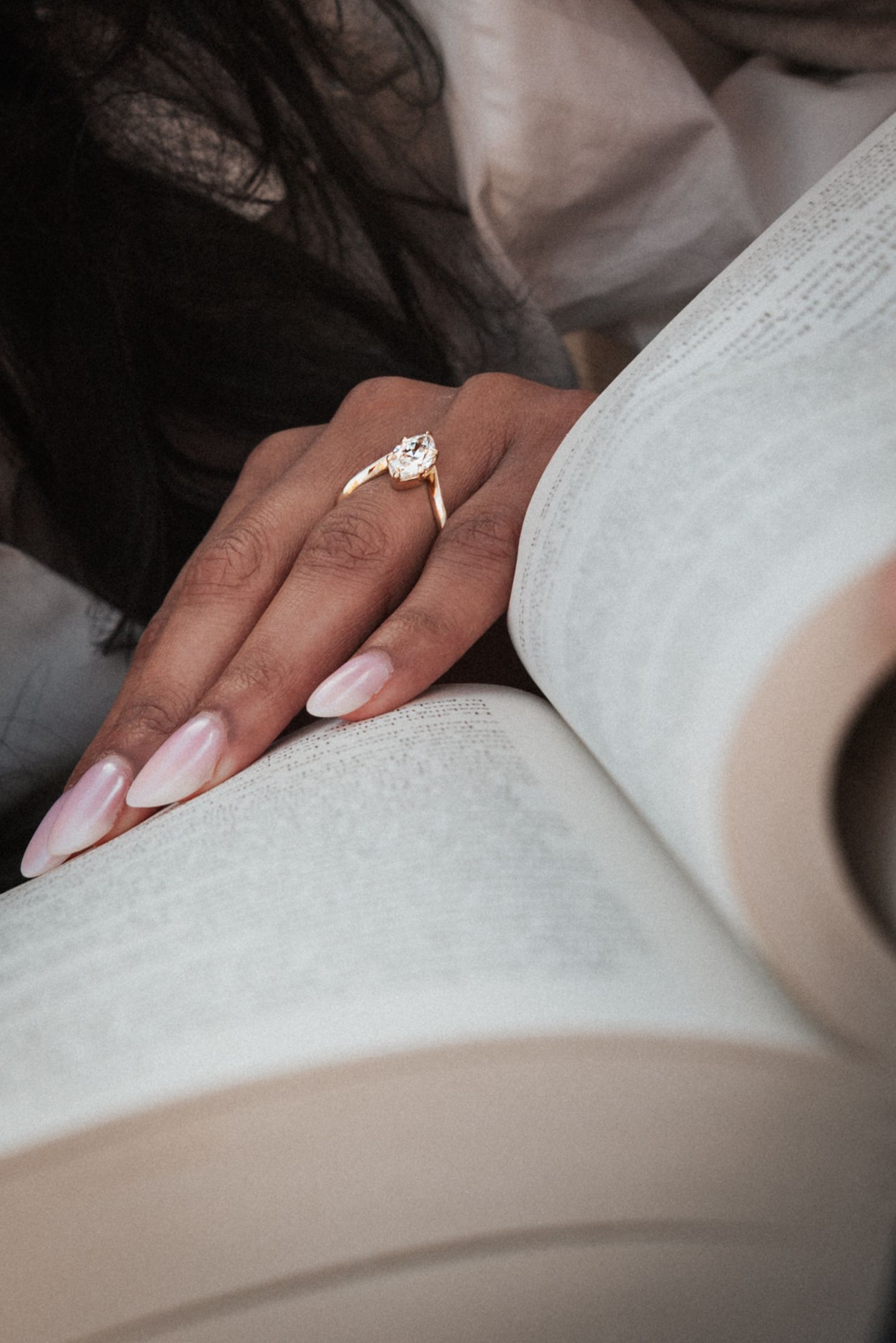 A hand with pink nails and a gold diamond engagement ring resting on an open book.