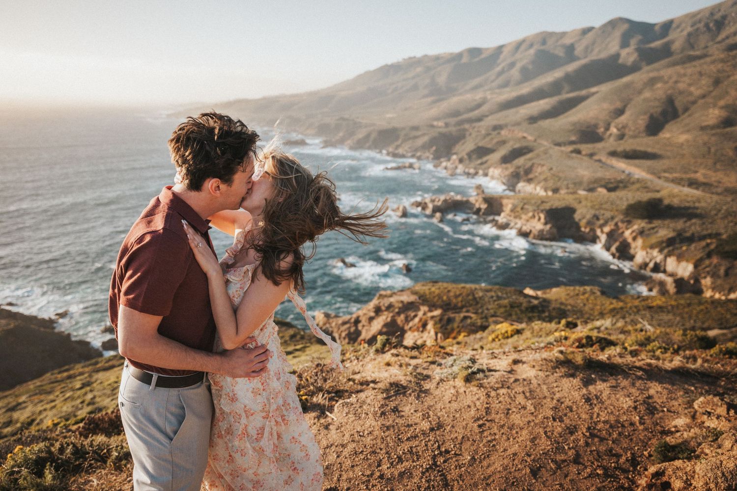 Couple kissing on a scenic Big Sur coastal cliffside with ocean waves and mountains in the golden sunset light.