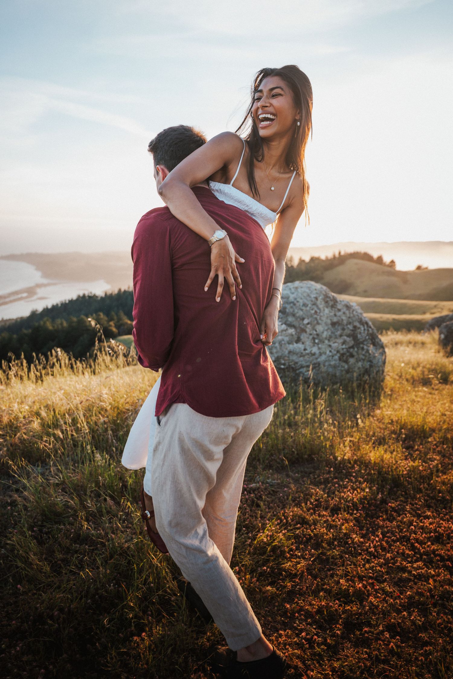 Couple embracing on a hillside at golden hour, woman laughing joyfully while being lifted by her partner.