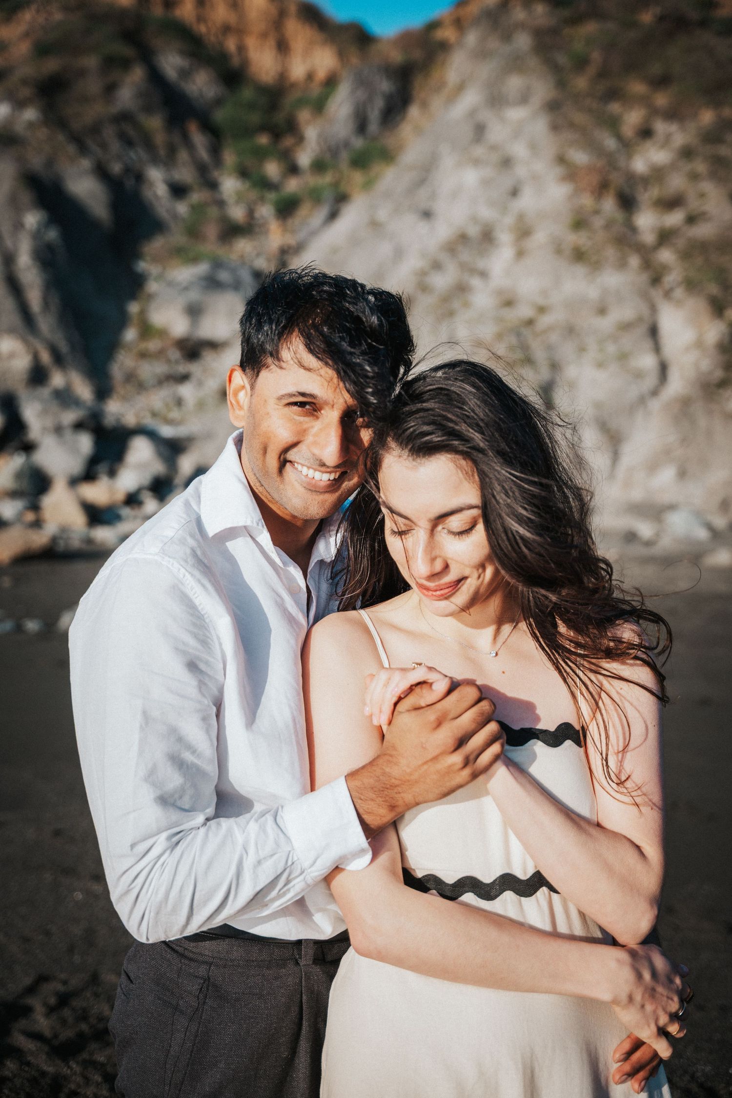 Romantic couple embracing on a rocky beach at sunset, man in white shirt hugging woman in white dress.