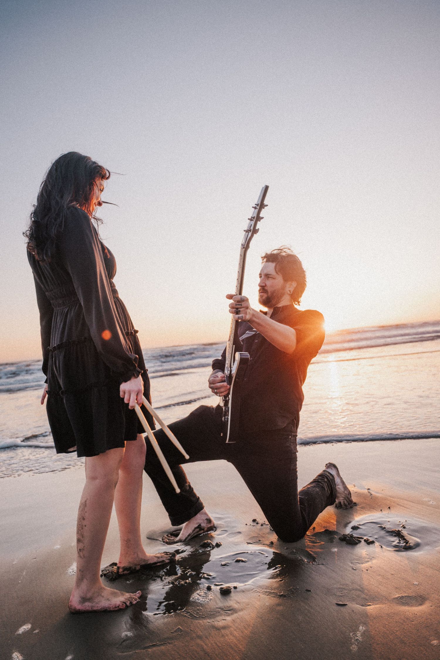 Man kneeling on beach at sunset playing guitar for woman in black dress, romantic ocean scene.