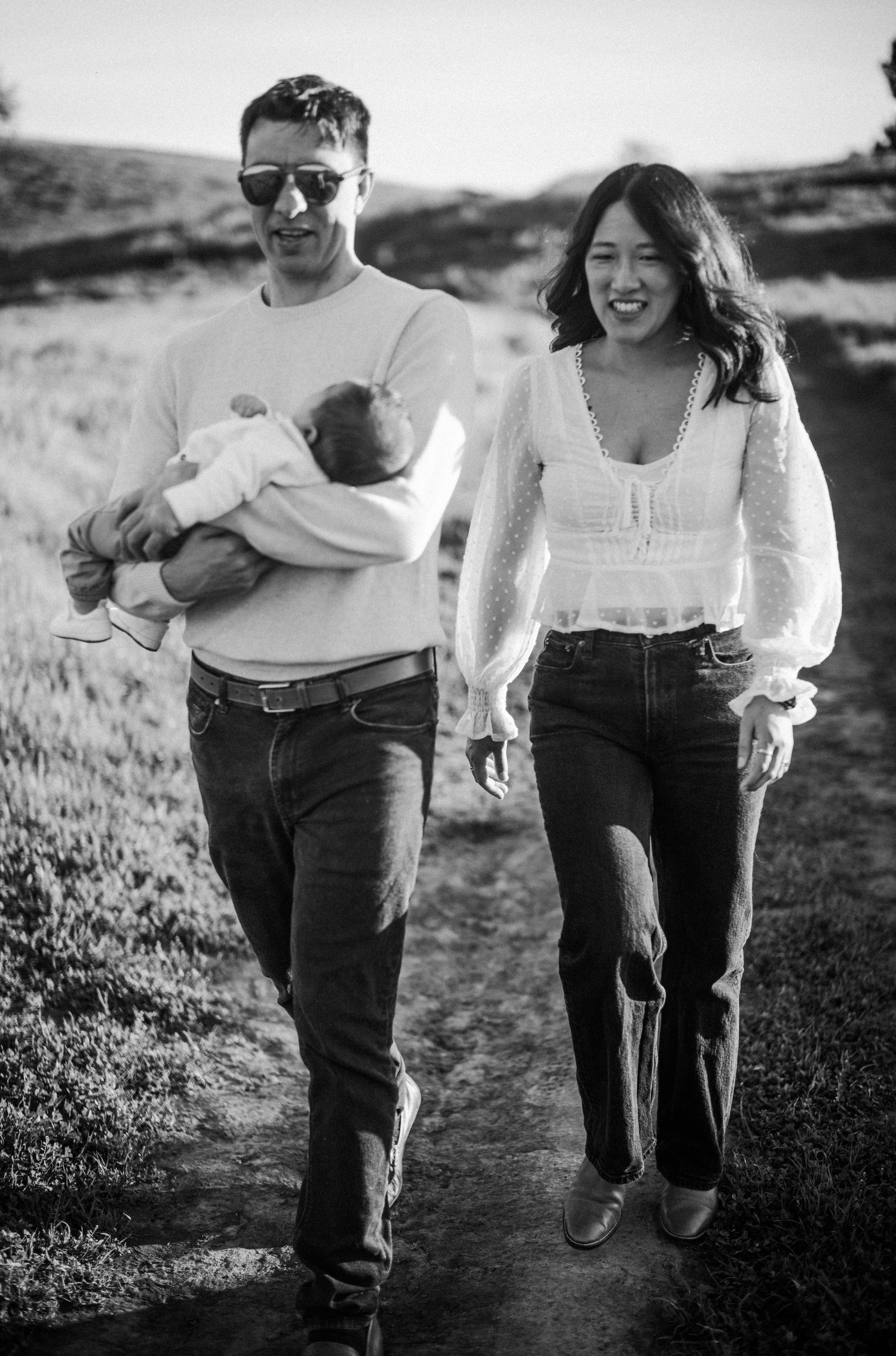Black and white photo of a couple walking outdoors, man holding a baby, woman smiling beside him.