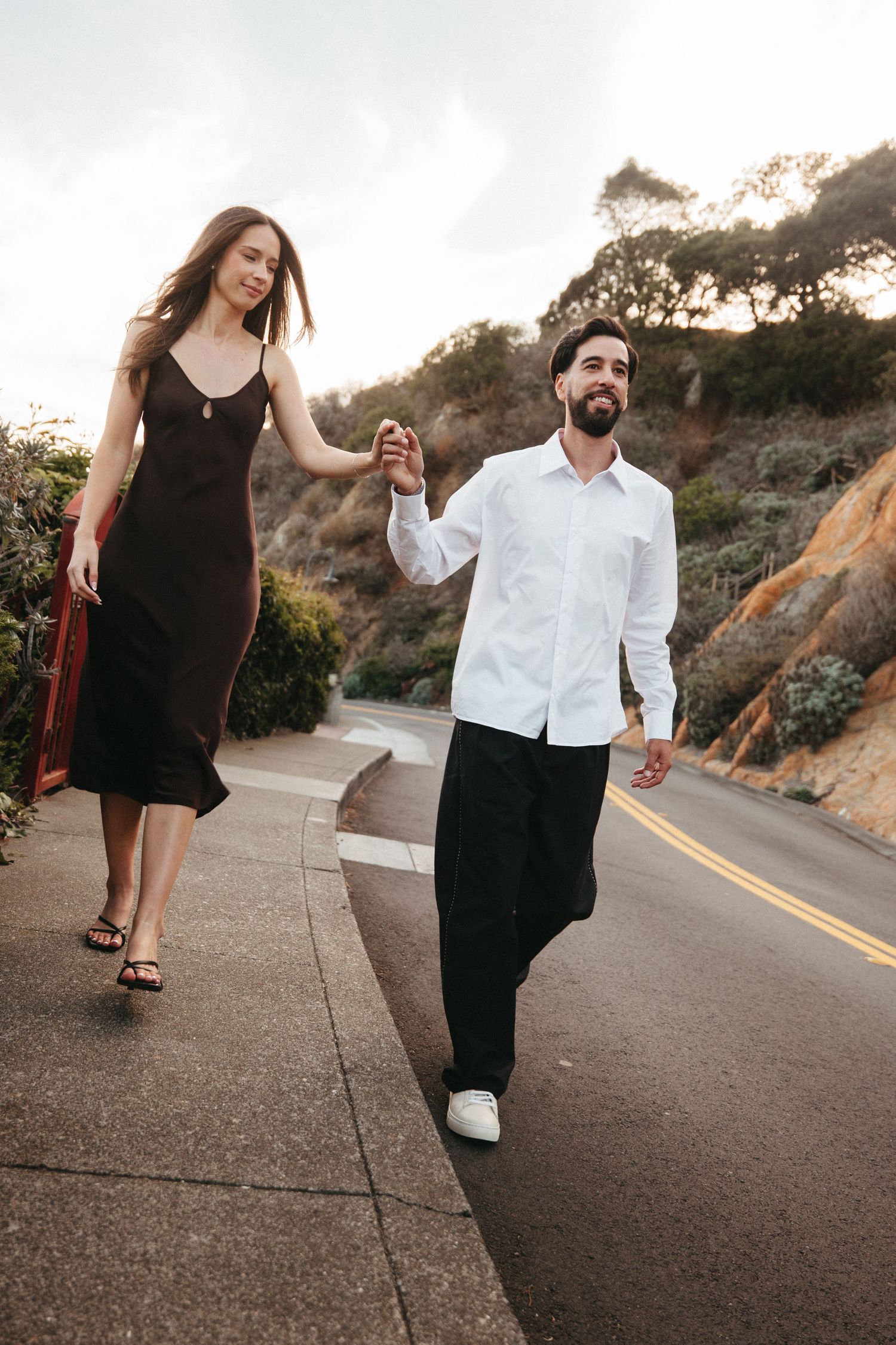 Happy couple holding hands while walking along a scenic road, woman in black dress and man in white shirt.