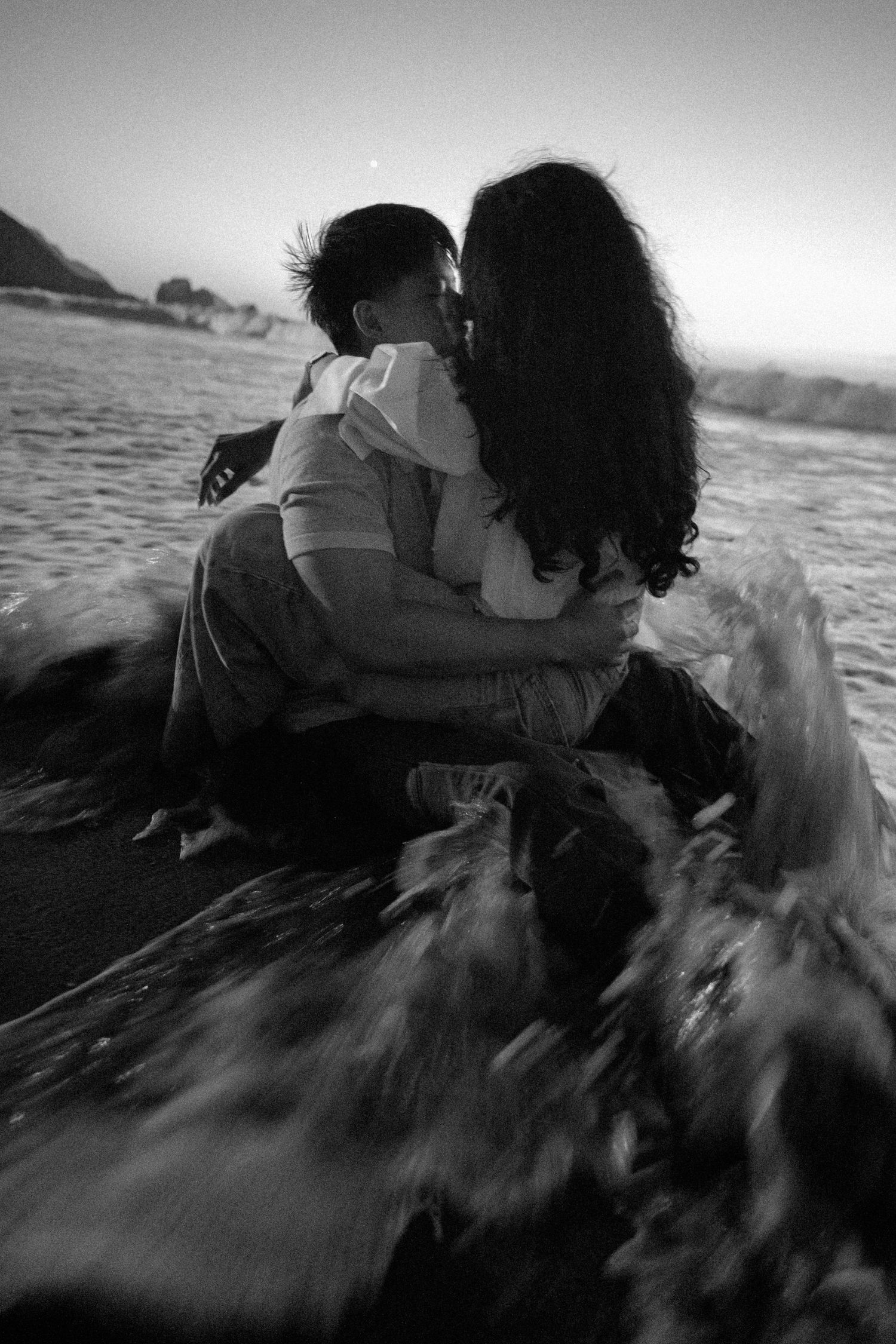 Black and white photo of a romantic couple kissing on a beach as ocean waves crash around them.