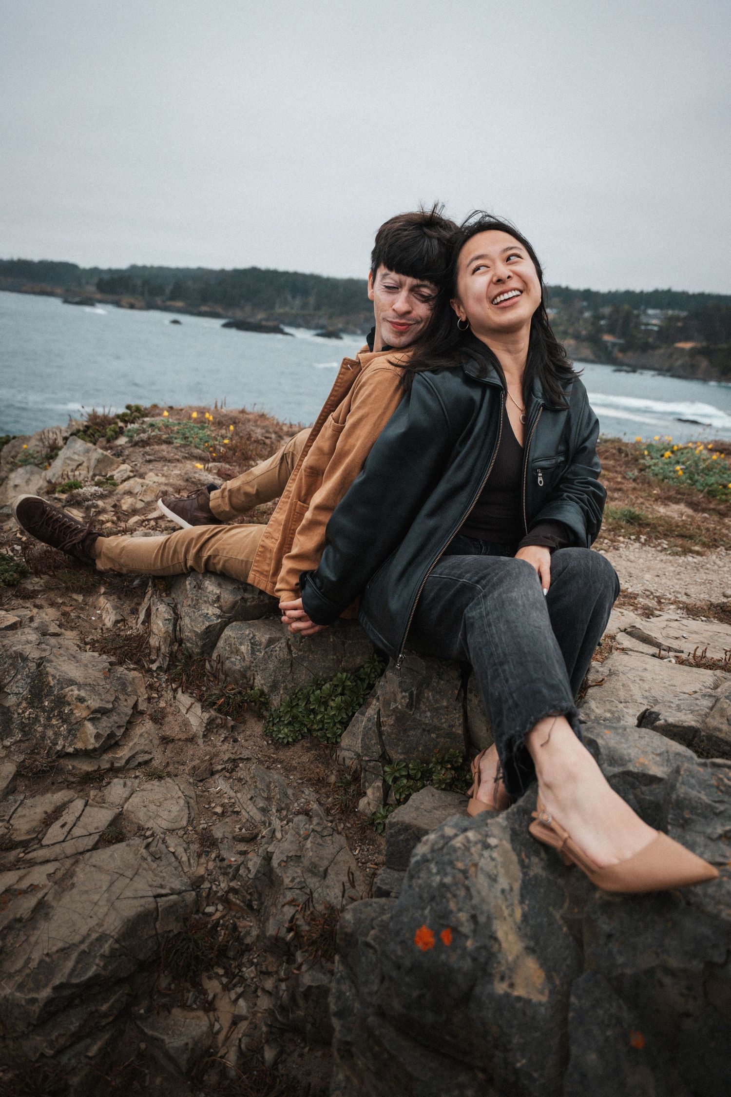Two women sitting on coastal rocks, laughing and holding hands with ocean views behind them.