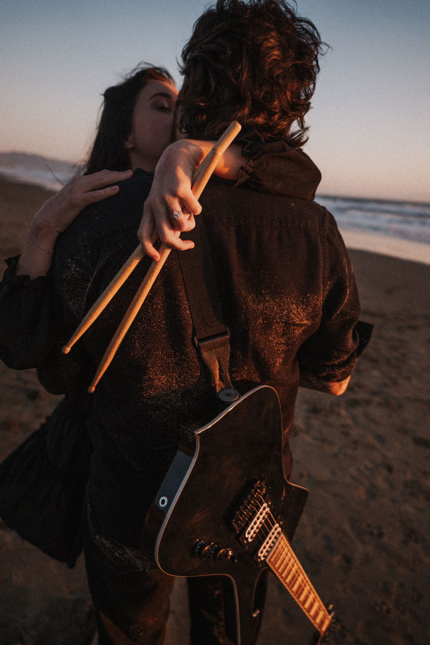 Couple embracing on beach at sunset, woman holding drumsticks, man carrying electric guitar.