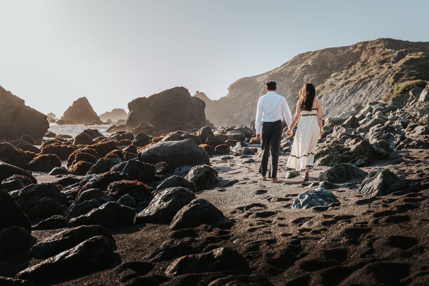 Couple holding hands walking on rocky beach toward ocean with dramatic cliffs in golden sunlight.