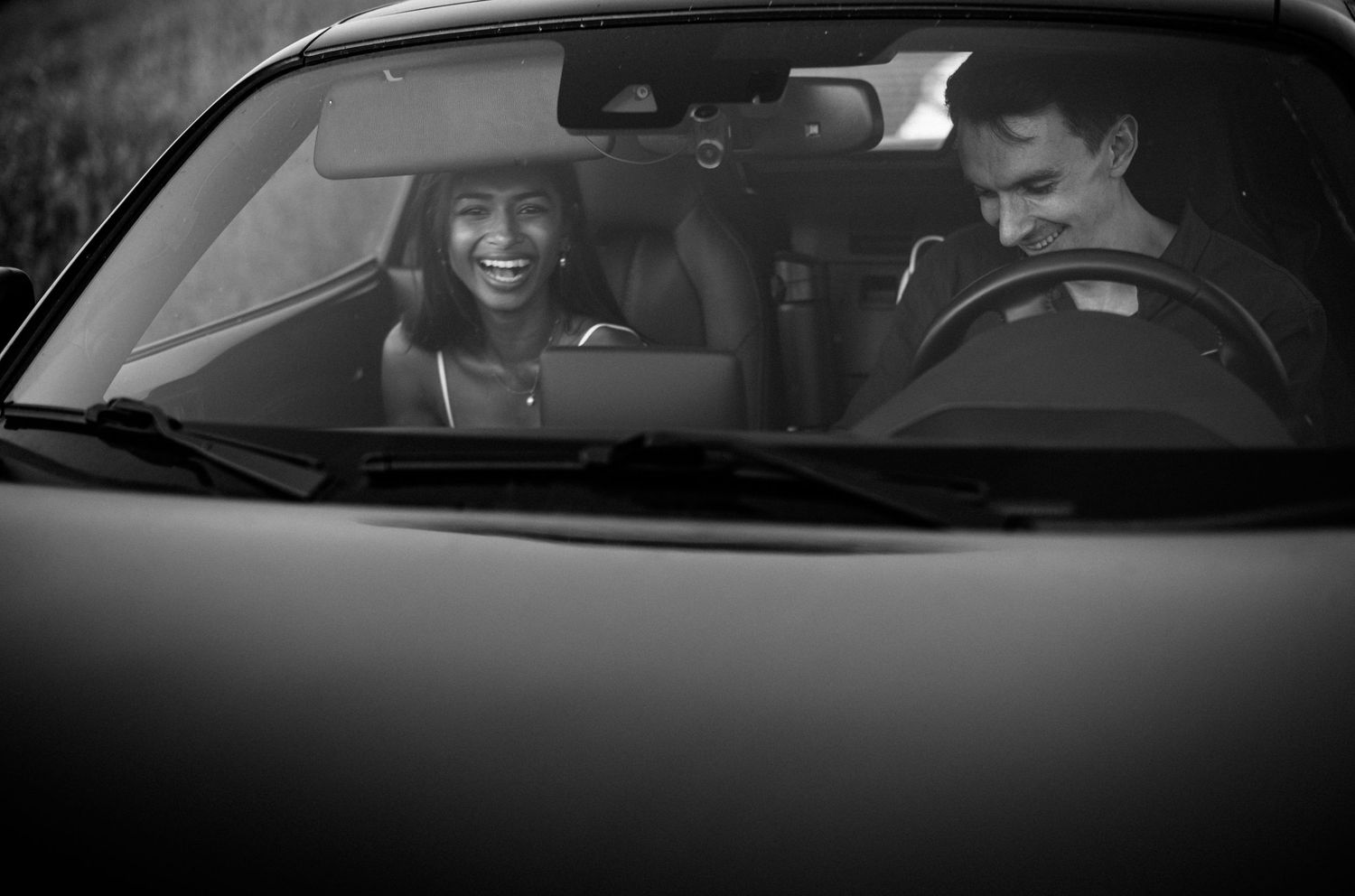 Black and white photo of a smiling couple sitting in a convertible car, enjoying a joyful moment together.