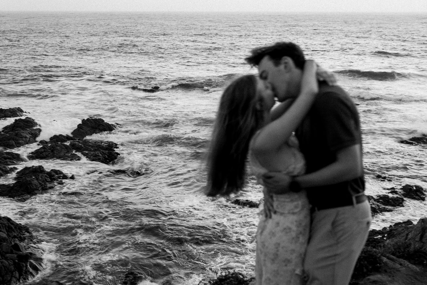Black and white photo of couple kissing passionately on rocky ocean shoreline.