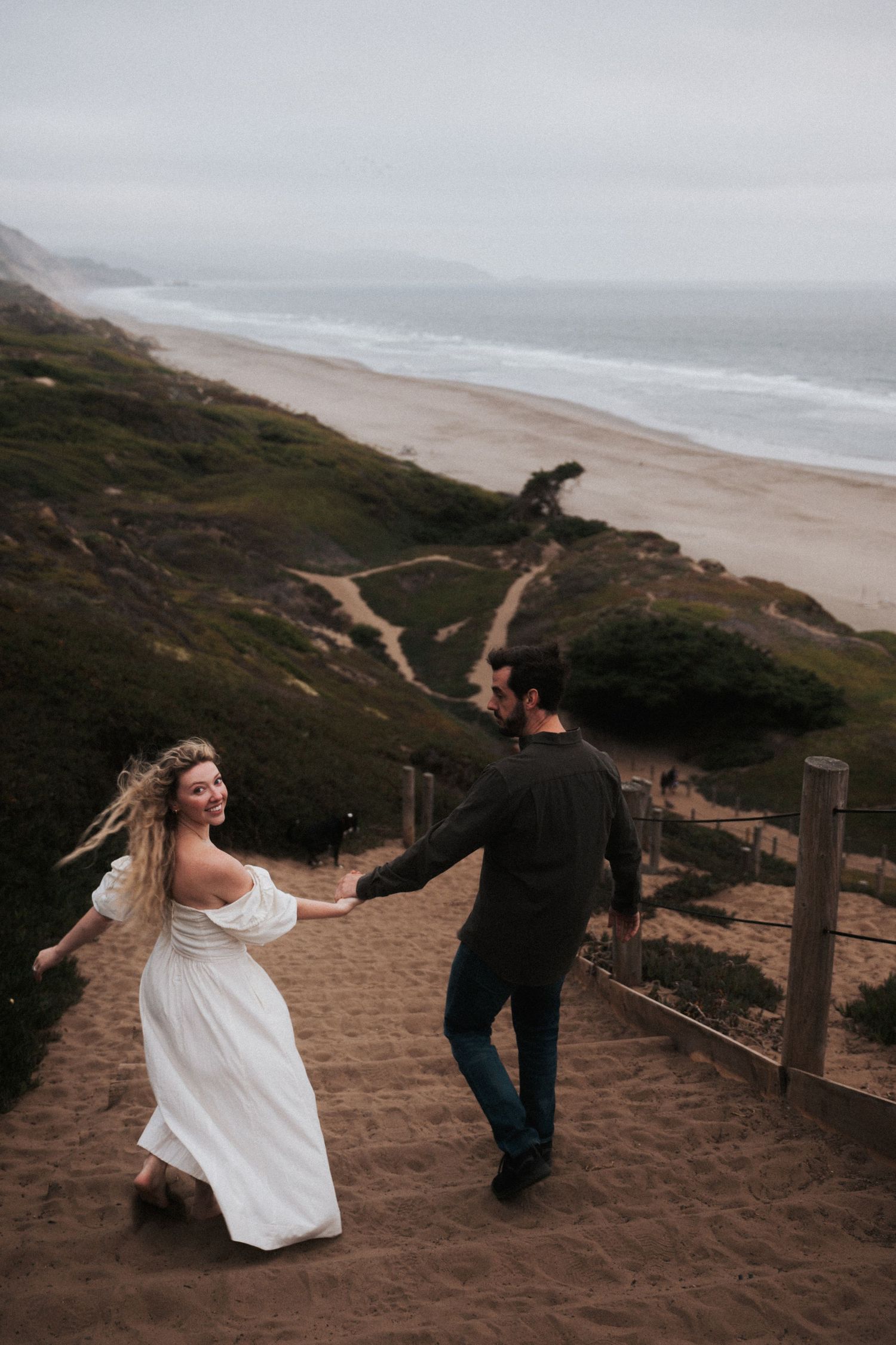 Couple holding hands walking down sandy beach stairs with ocean views on cloudy day.