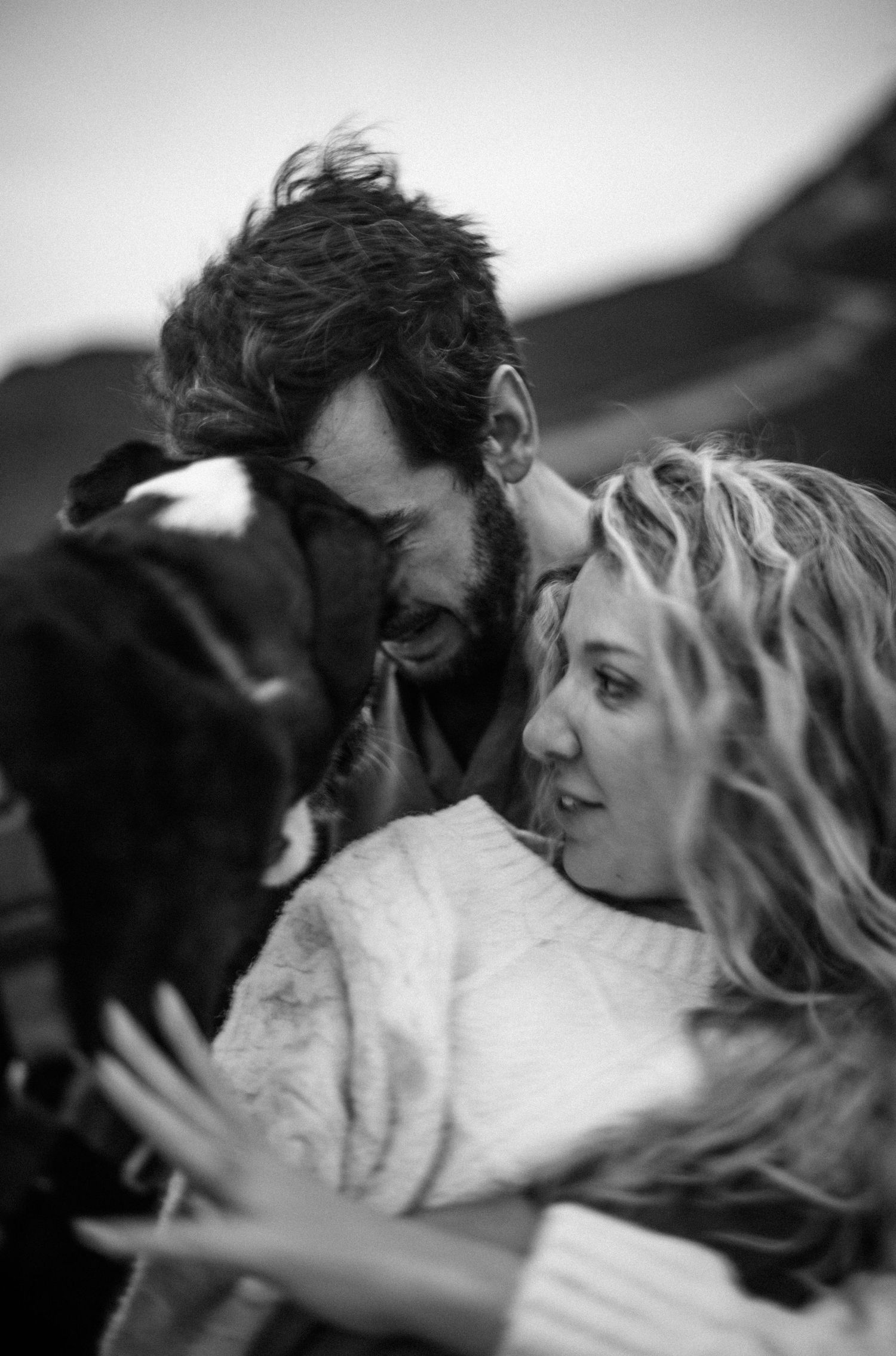 Black and white photo of a couple smiling and cuddling with their dog outdoors.