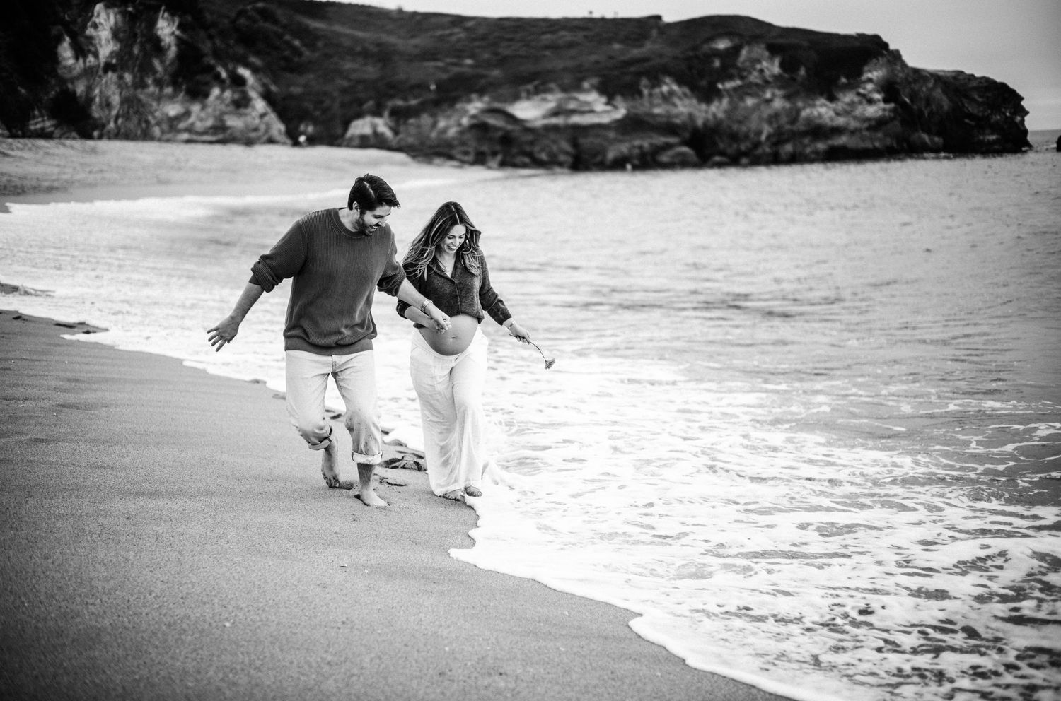 Couple walking along sandy beach shore in black and white photo with rocky cliffs in background.