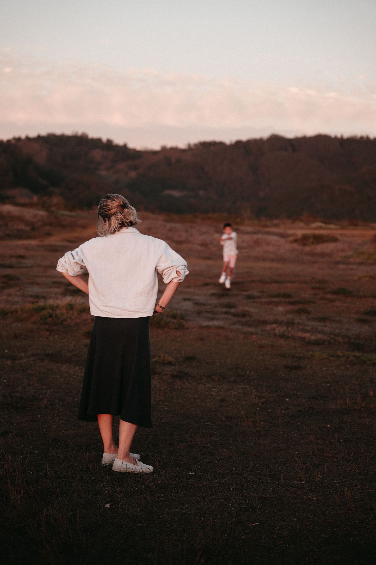 Two women standing in a vast open field at dusk, one in a white jacket and black skirt facing away.