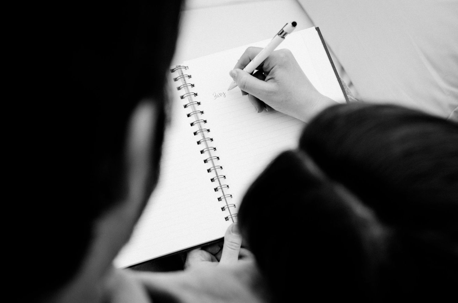 Black and white photo of a person writing in a spiral notebook with a pencil, viewed from above.