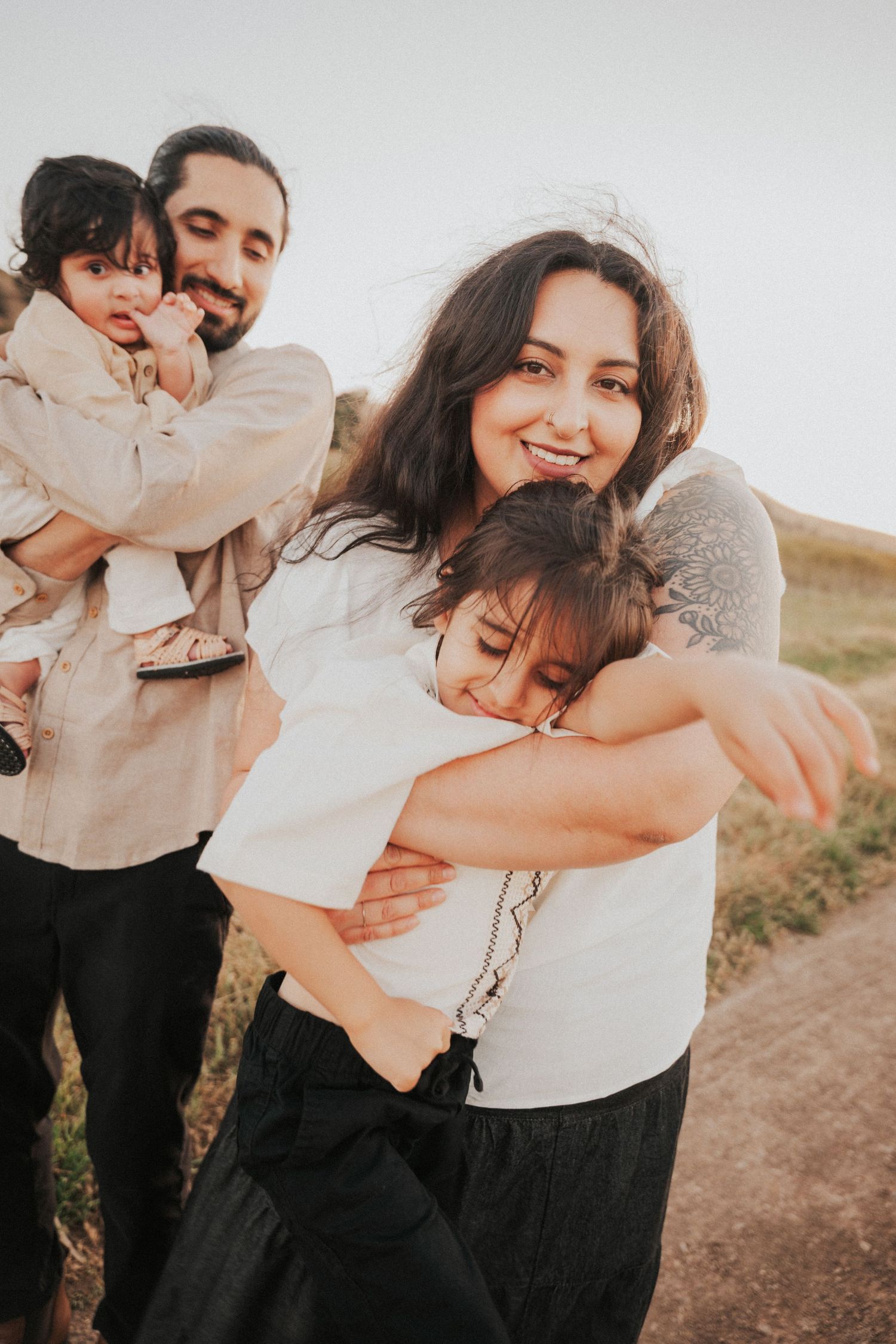 A happy family embraces outdoors, mother hugging child while father holds baby in golden field light.