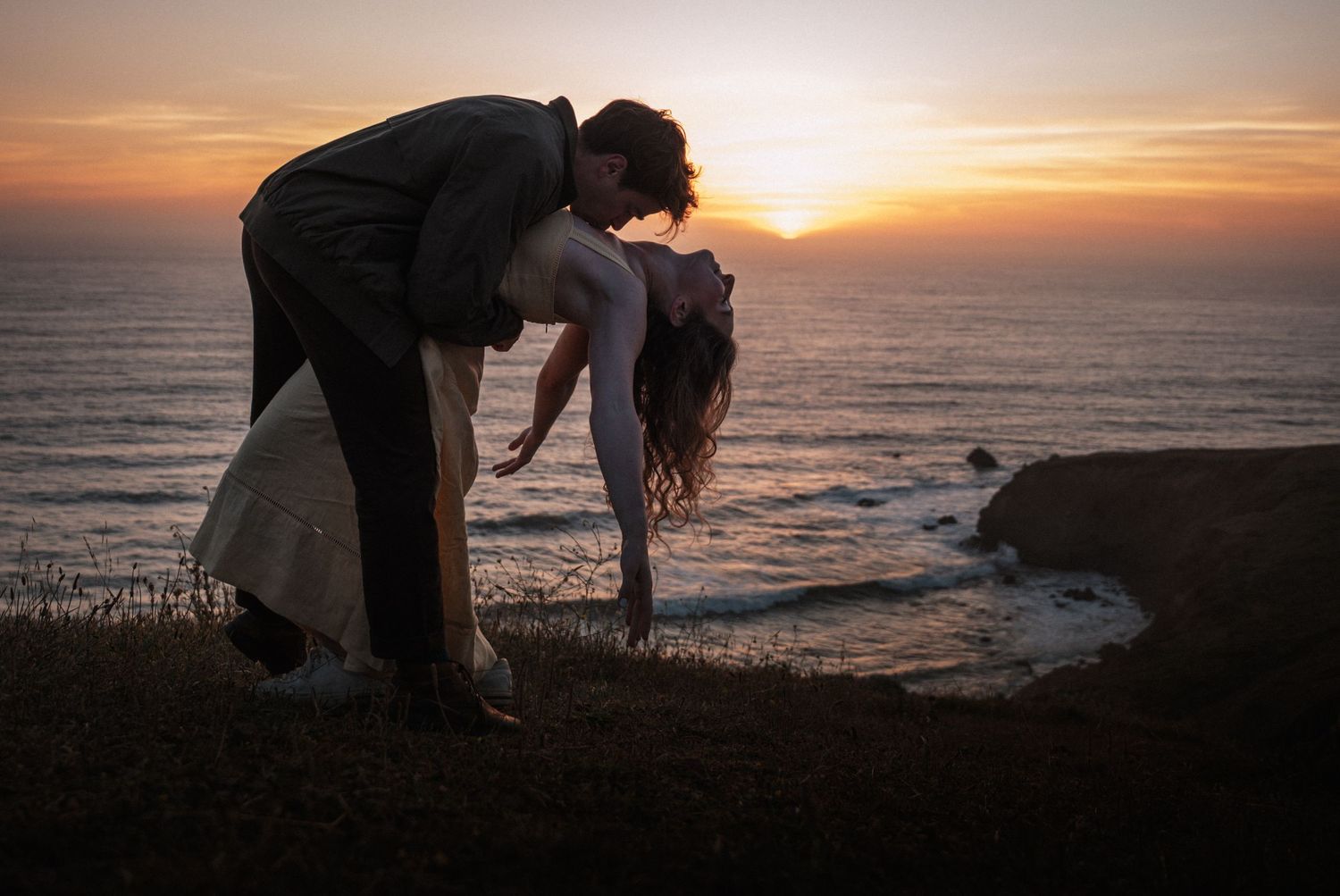 Couple sharing a romantic dip kiss silhouetted against a stunning ocean sunset on a coastal cliff.