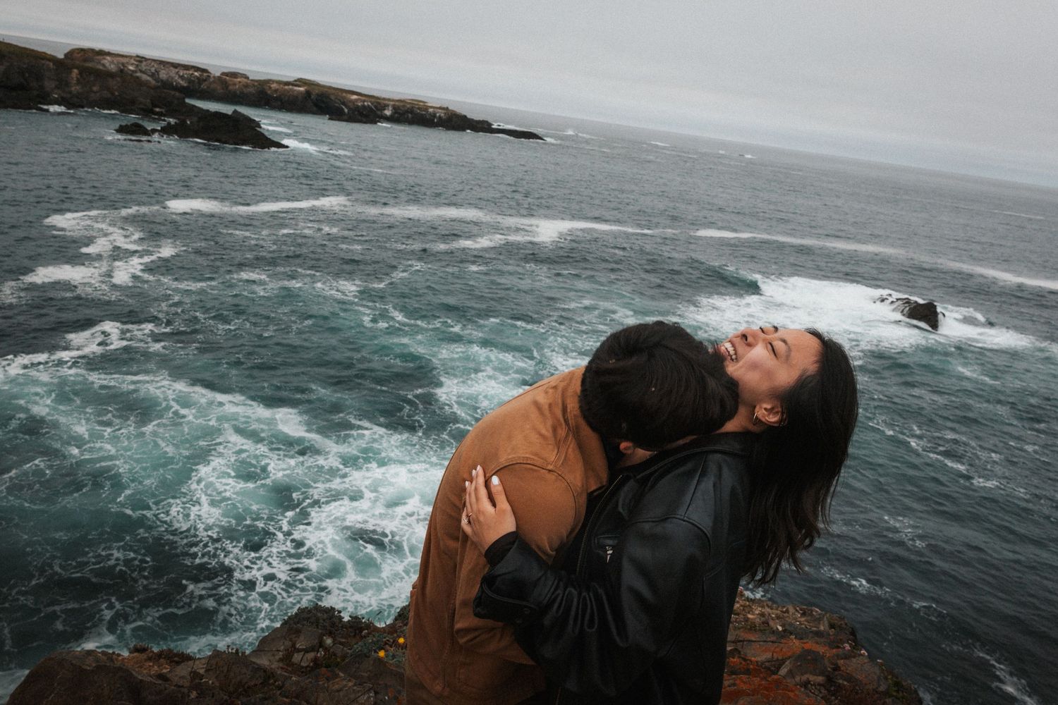 Romantic couple embracing on rocky coastal cliffs with dramatic ocean waves crashing in the background.