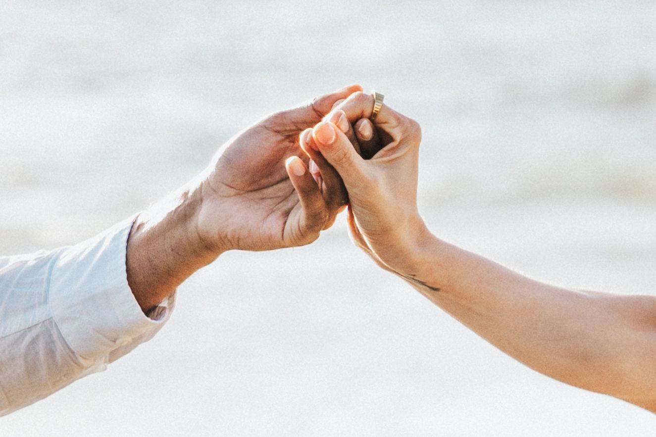 Close-up of two hands gently holding fingers together on a blurred beach background.