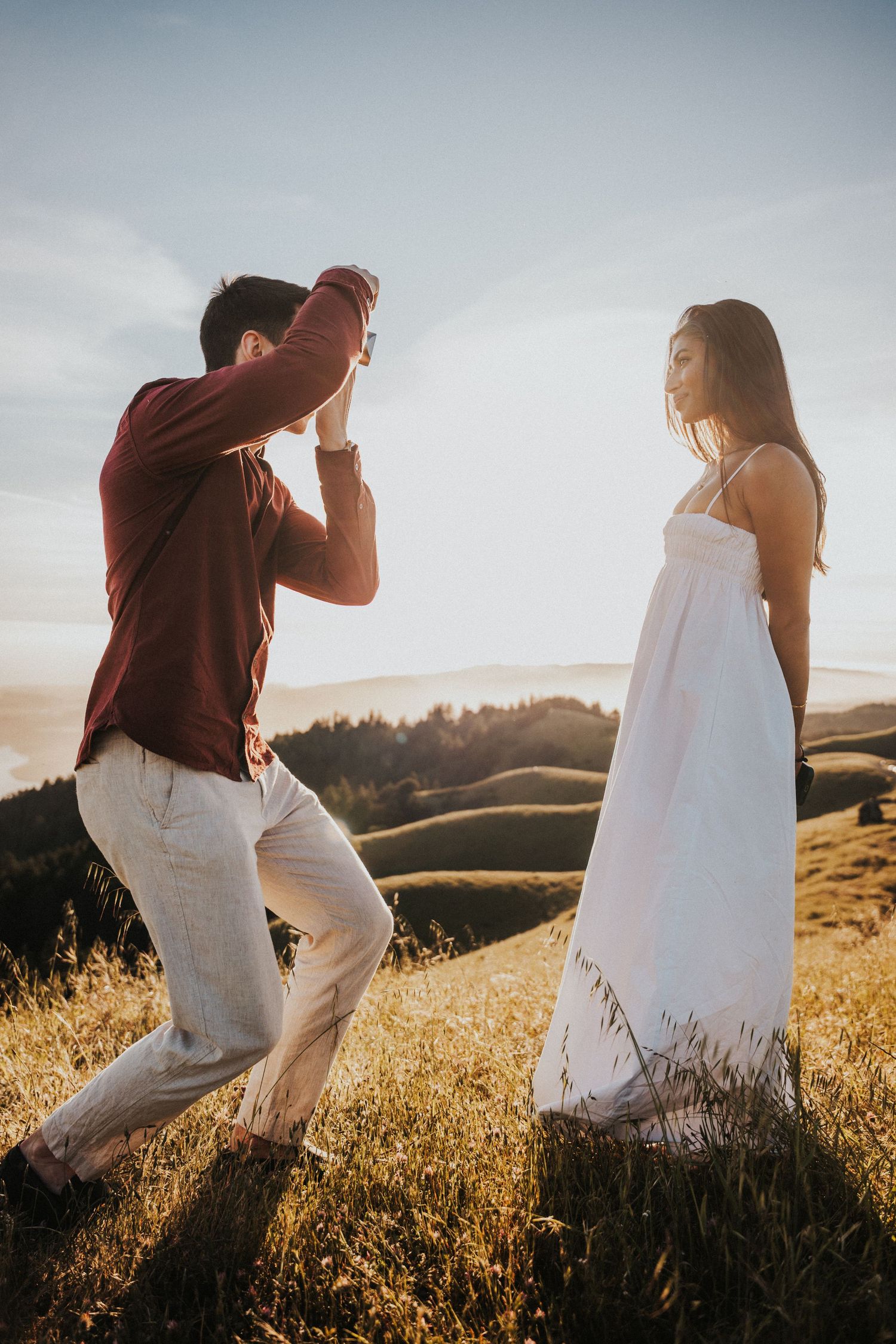 Man photographing woman in white dress on golden hillside at sunset with scenic valley views.Photography by The Y.K. Studio.