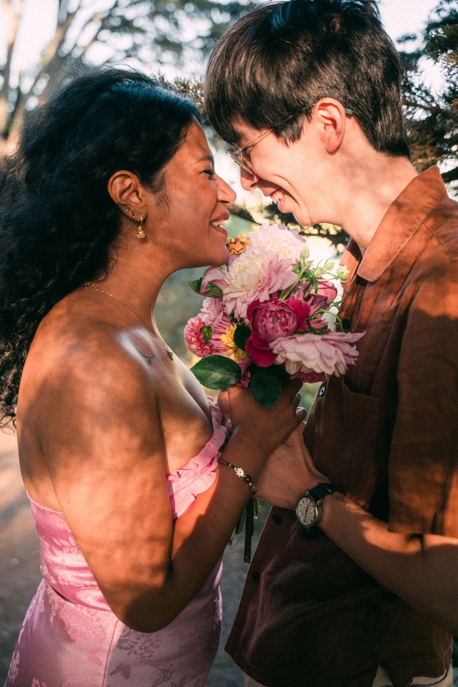 Laughing couple sharing a bouquet of pink peonies in warm golden sunlight outdoors.