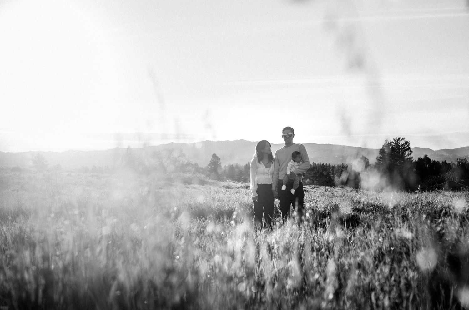 Black and white photo of a couple standing together in a sunlit meadow field with mountains in background.