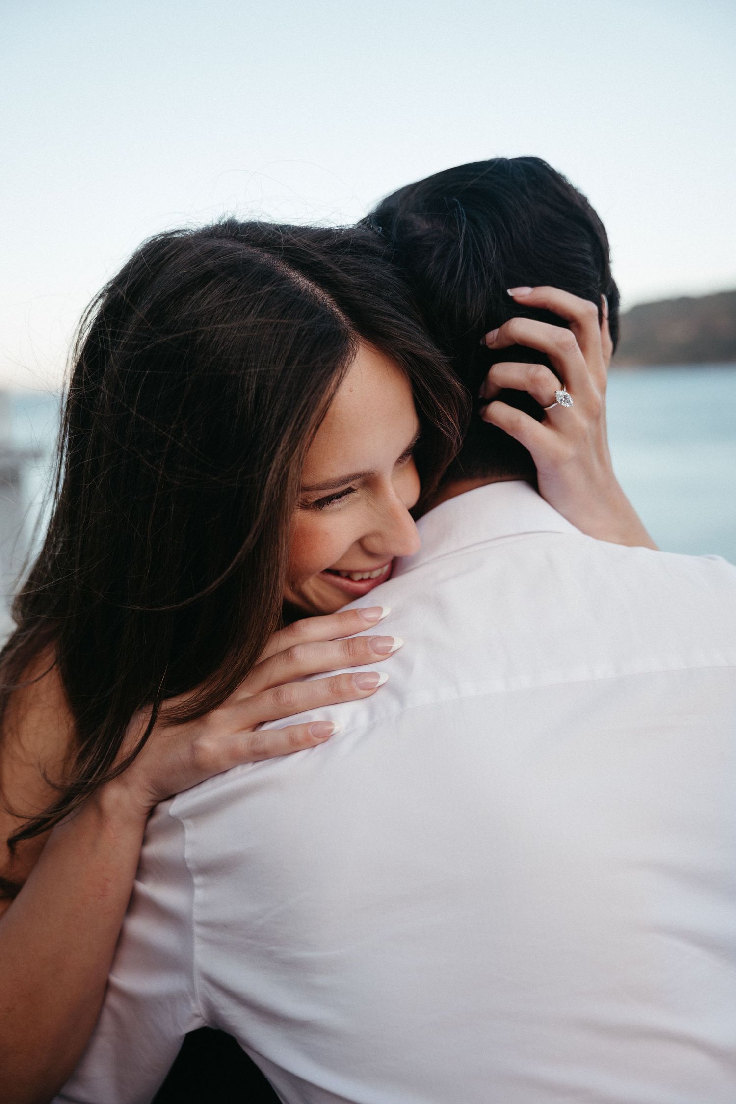 A smiling woman with an engagement ring embraces a man near the water in a romantic outdoor setting.