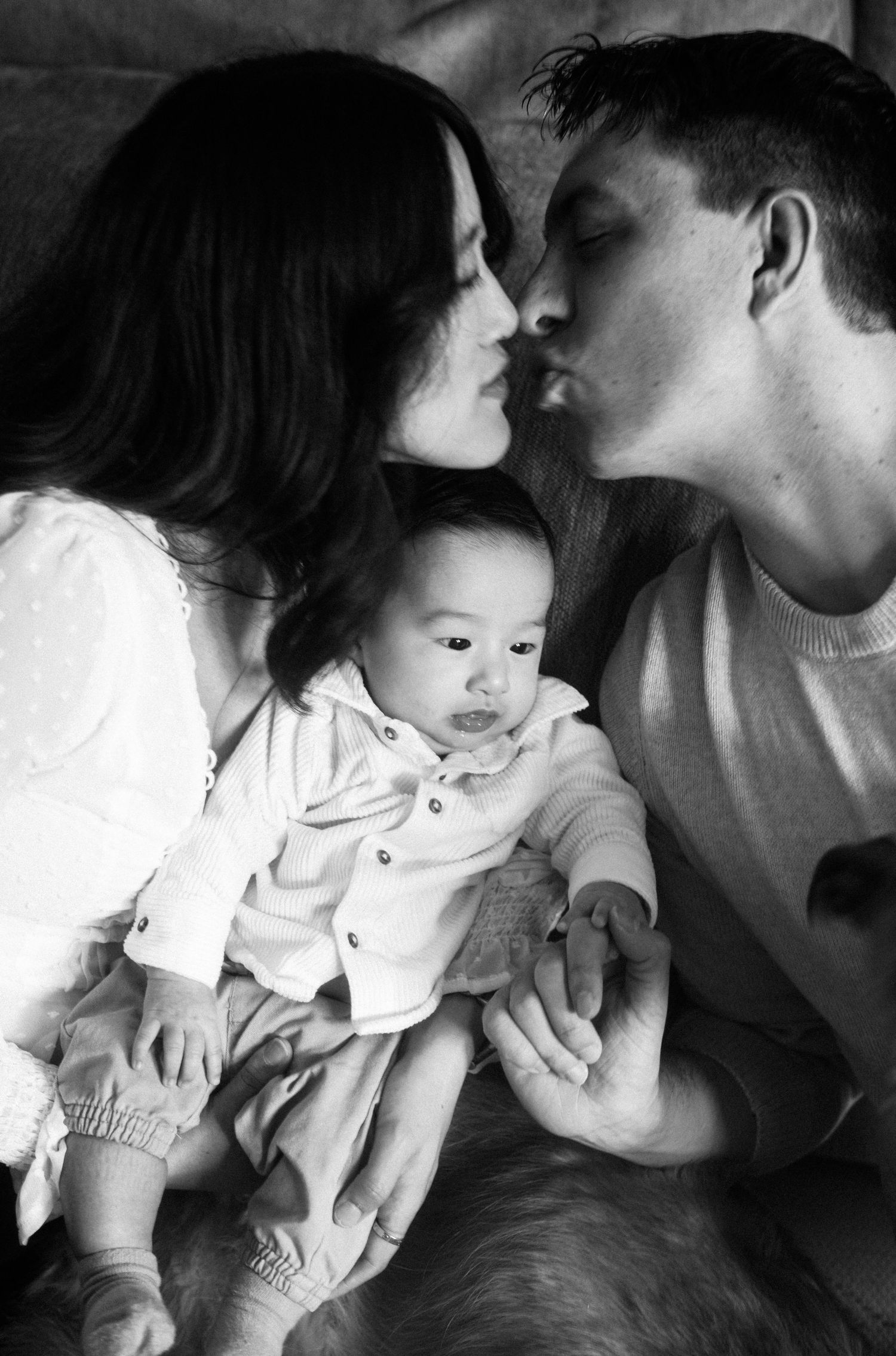 Black and white photo of parents kissing while holding their newborn baby between them.
