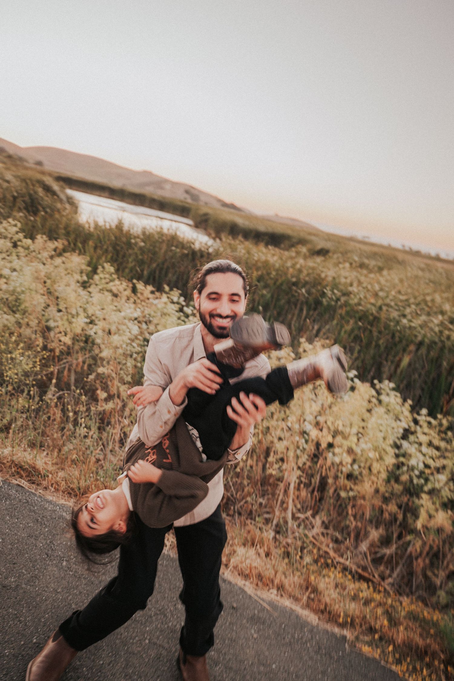 A bearded man joyfully lifts a laughing woman upside down on a scenic road surrounded by lush greenery and hills.