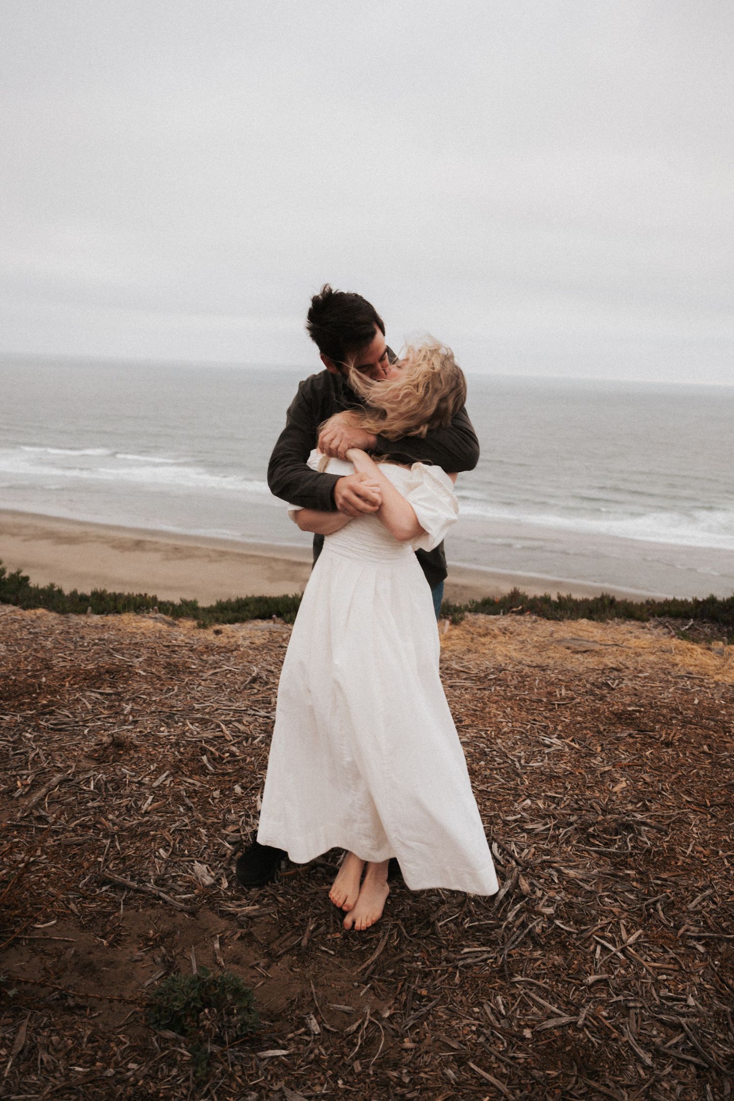 Couple embracing and kissing on a coastal bluff, woman in white dress, ocean waves in background on overcast day.