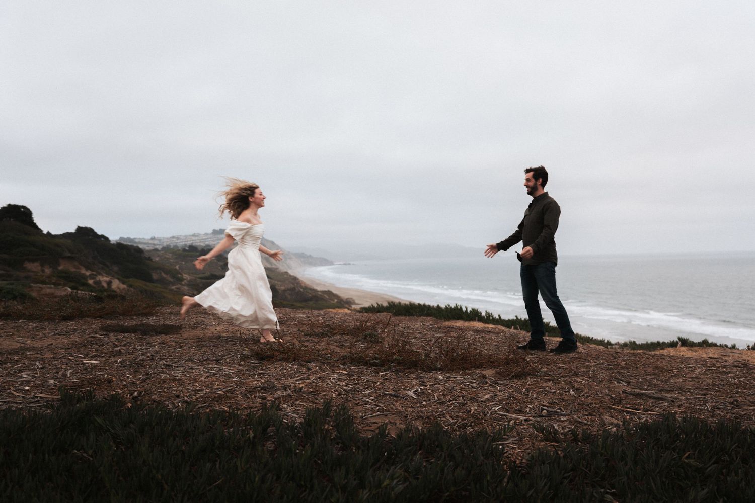 A bride in a white dress runs toward a groom on a coastal cliffside with ocean views on an overcast day.