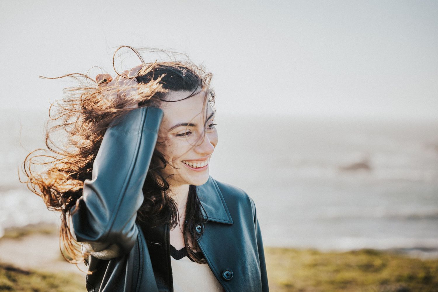 Smiling young woman in leather jacket with wind-blown hair near ocean shore.