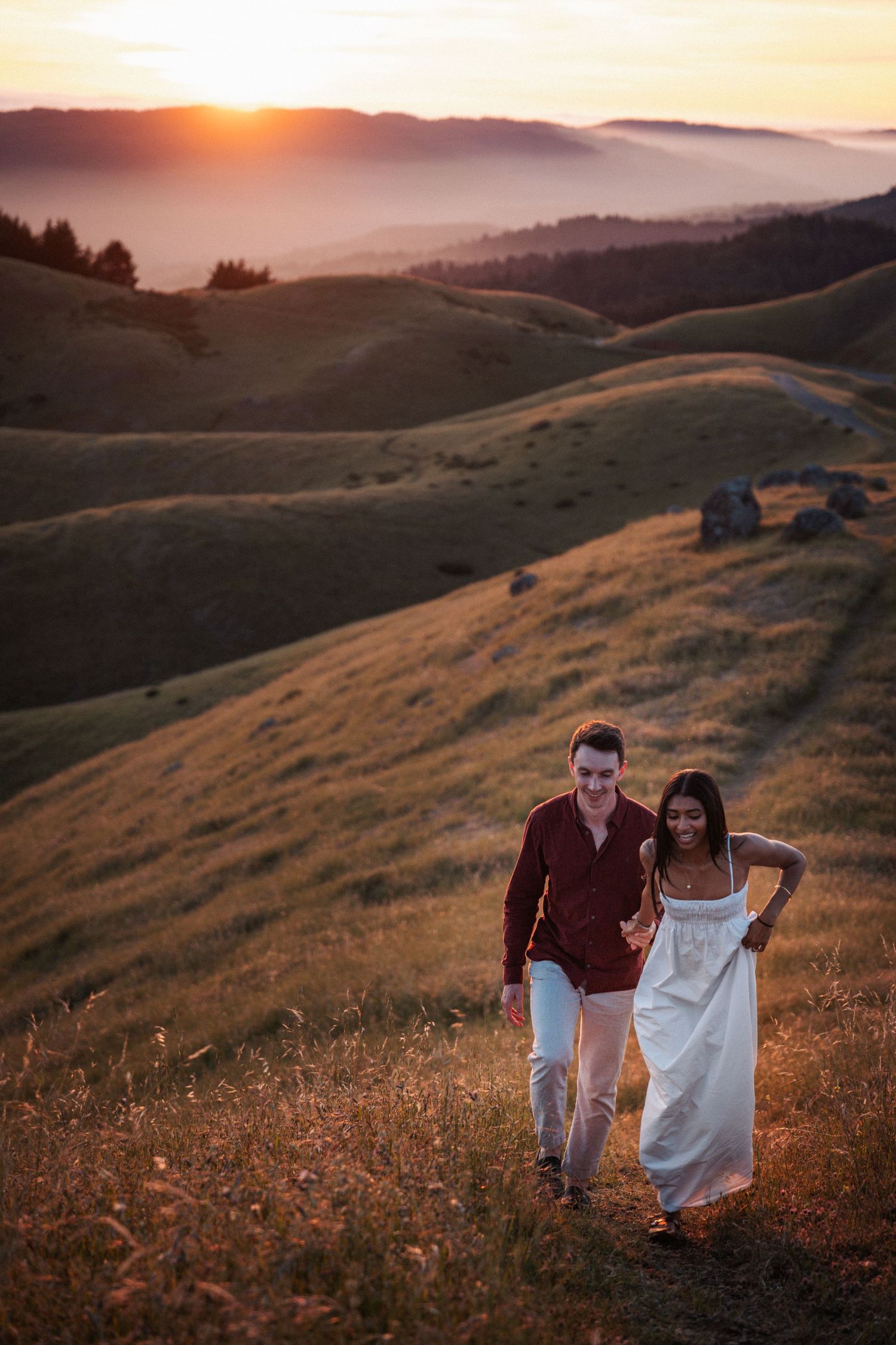 Couple walking hand in hand through a grassy hillside at golden sunset with misty mountains behind.