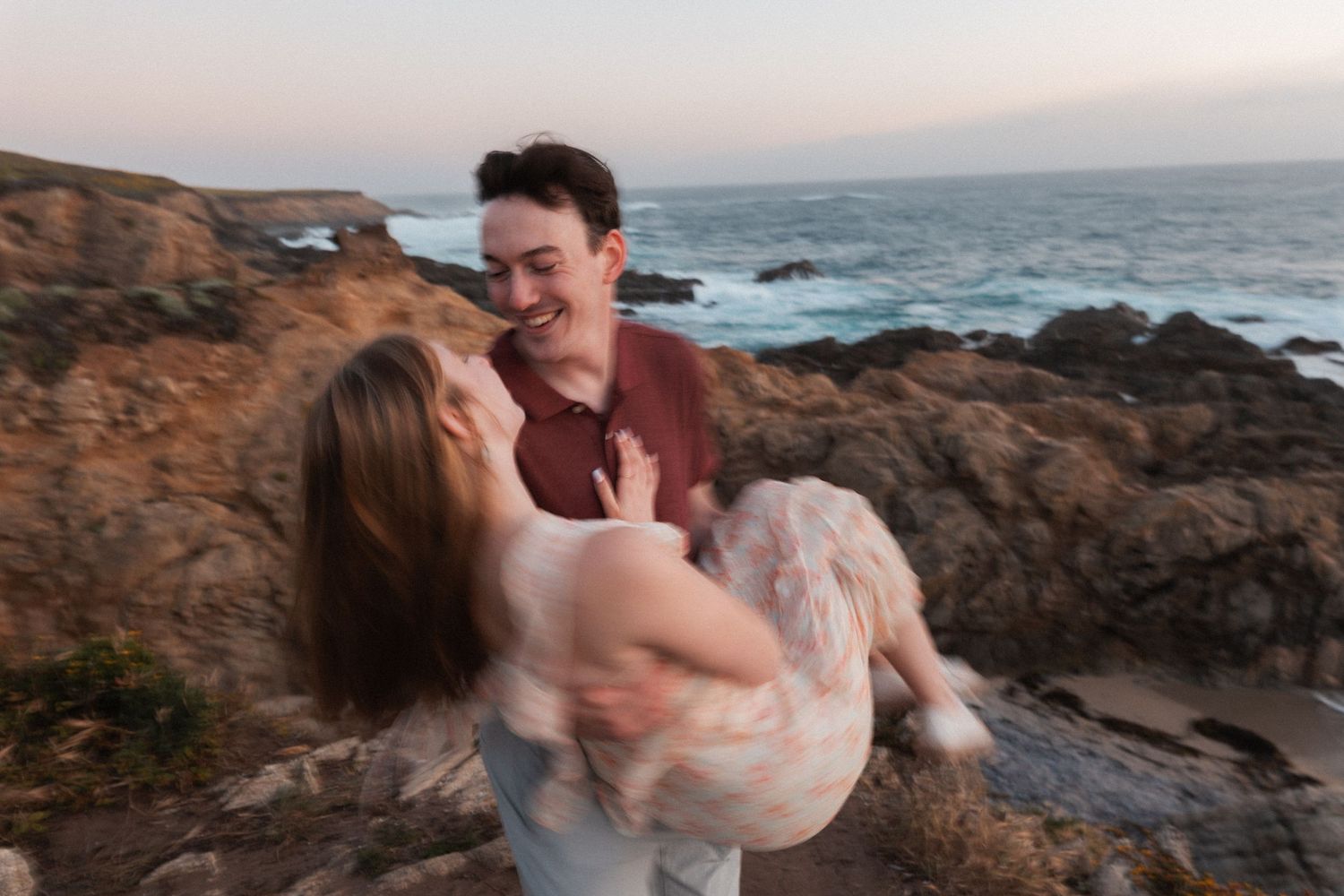 Man lifting woman near rocky ocean shore at dusk, both laughing joyfully.