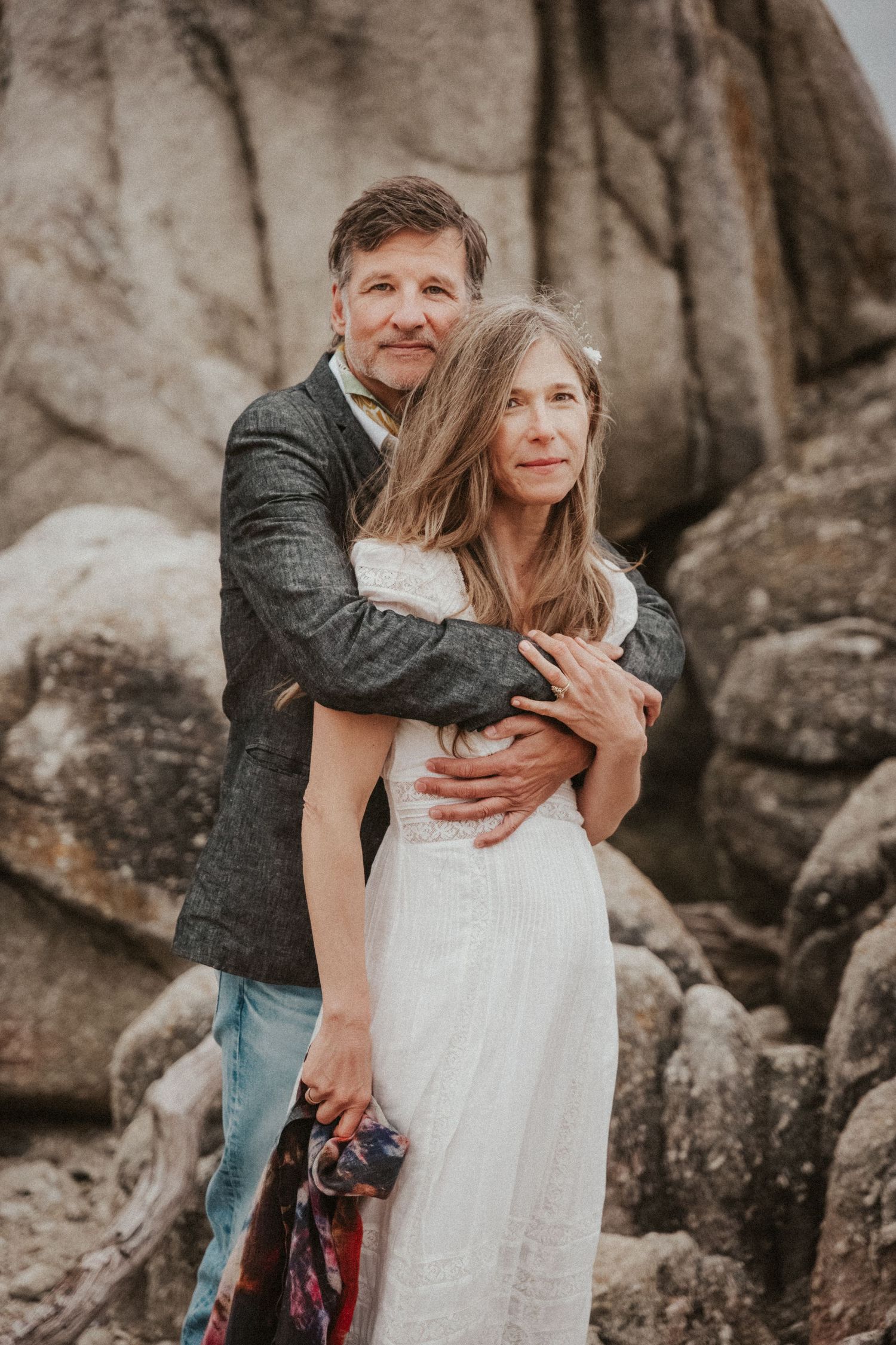 Man in gray jacket tenderly embraces woman in white wedding dress against dramatic rocky boulder background outdoors.