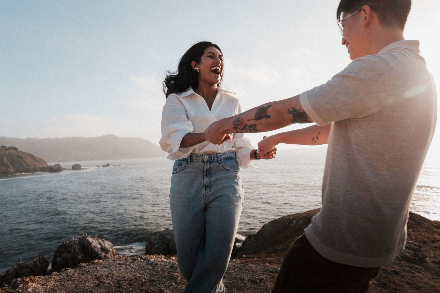 A joyful couple holds hands on a rocky coastal cliff at sunset, with ocean waves and mountains in the background.