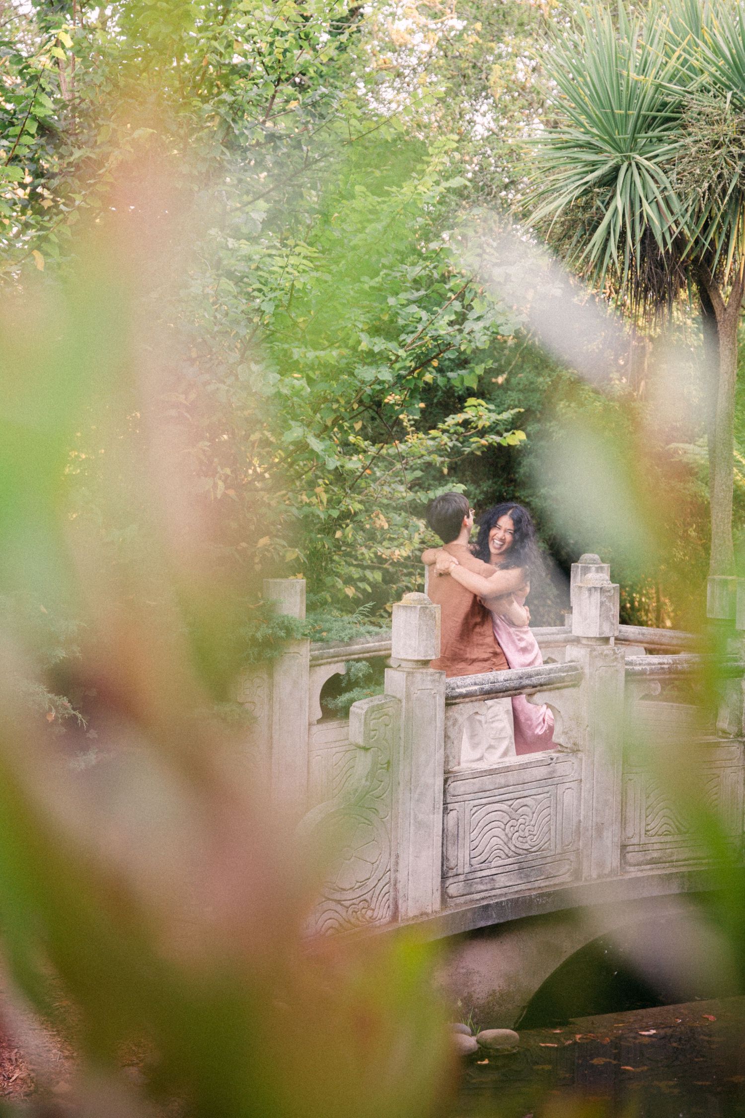 Couple embracing on an ornate stone bridge surrounded by lush green trees in a serene garden setting.